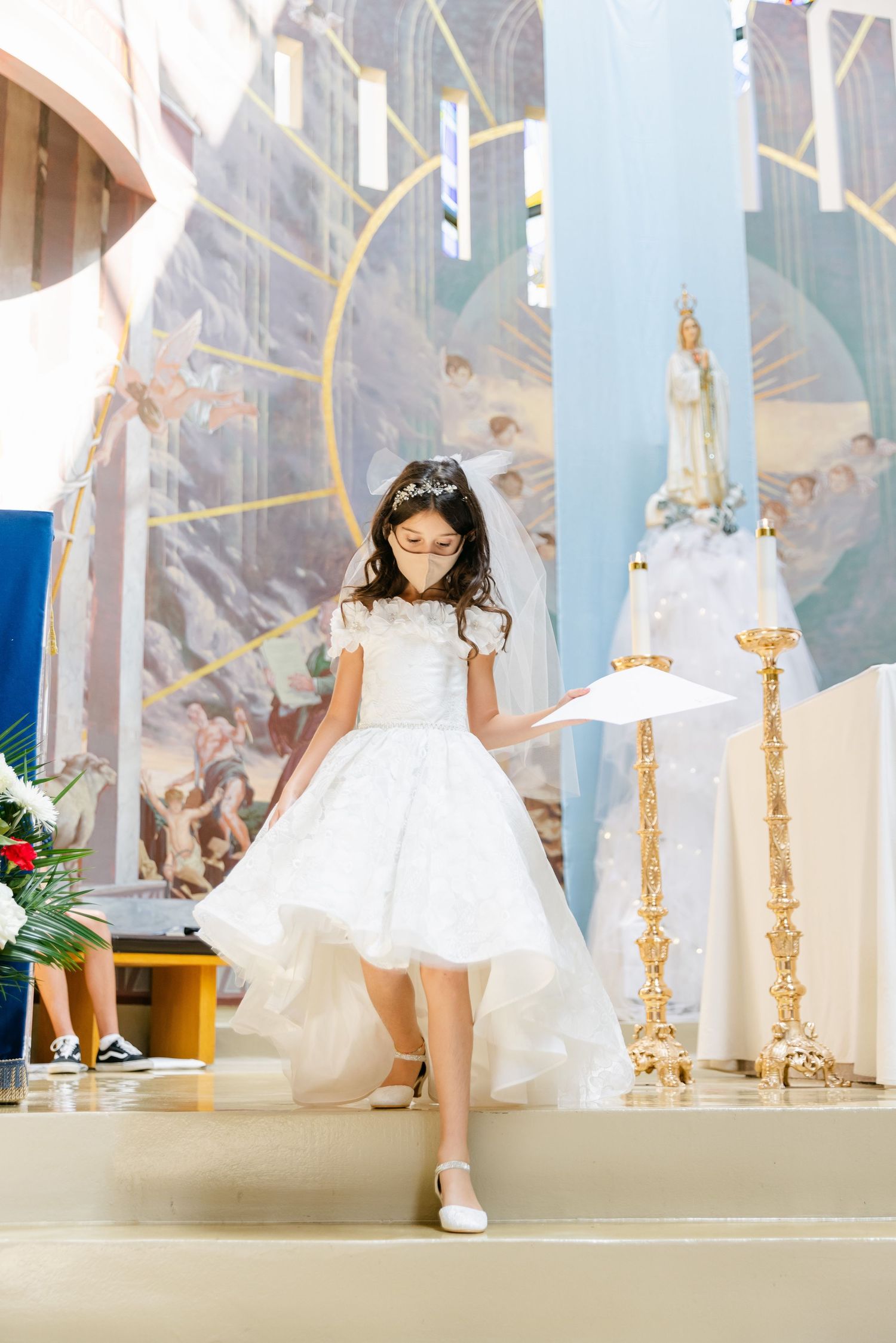 A young girl in a white dress and tiara poses in front of church murals and religious statues.