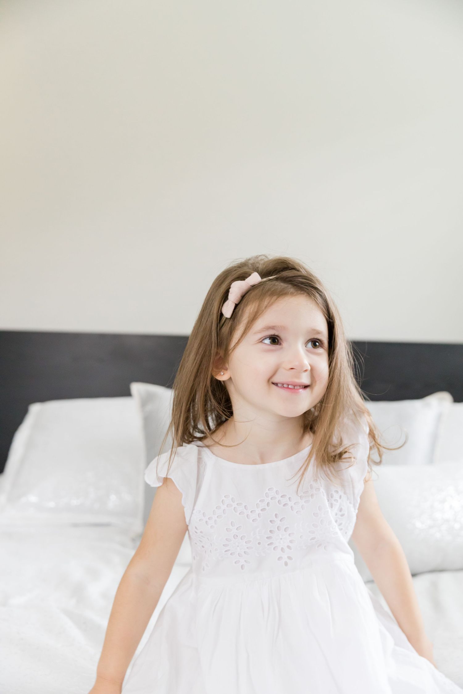 Child in white eyelet dress smiles while sitting on white bed.