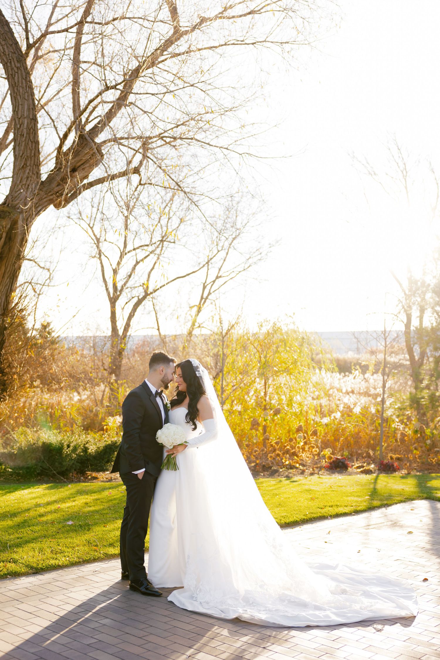 Wedding couple embraces on a brick walkway surrounded by autumn trees and warm afternoon light.