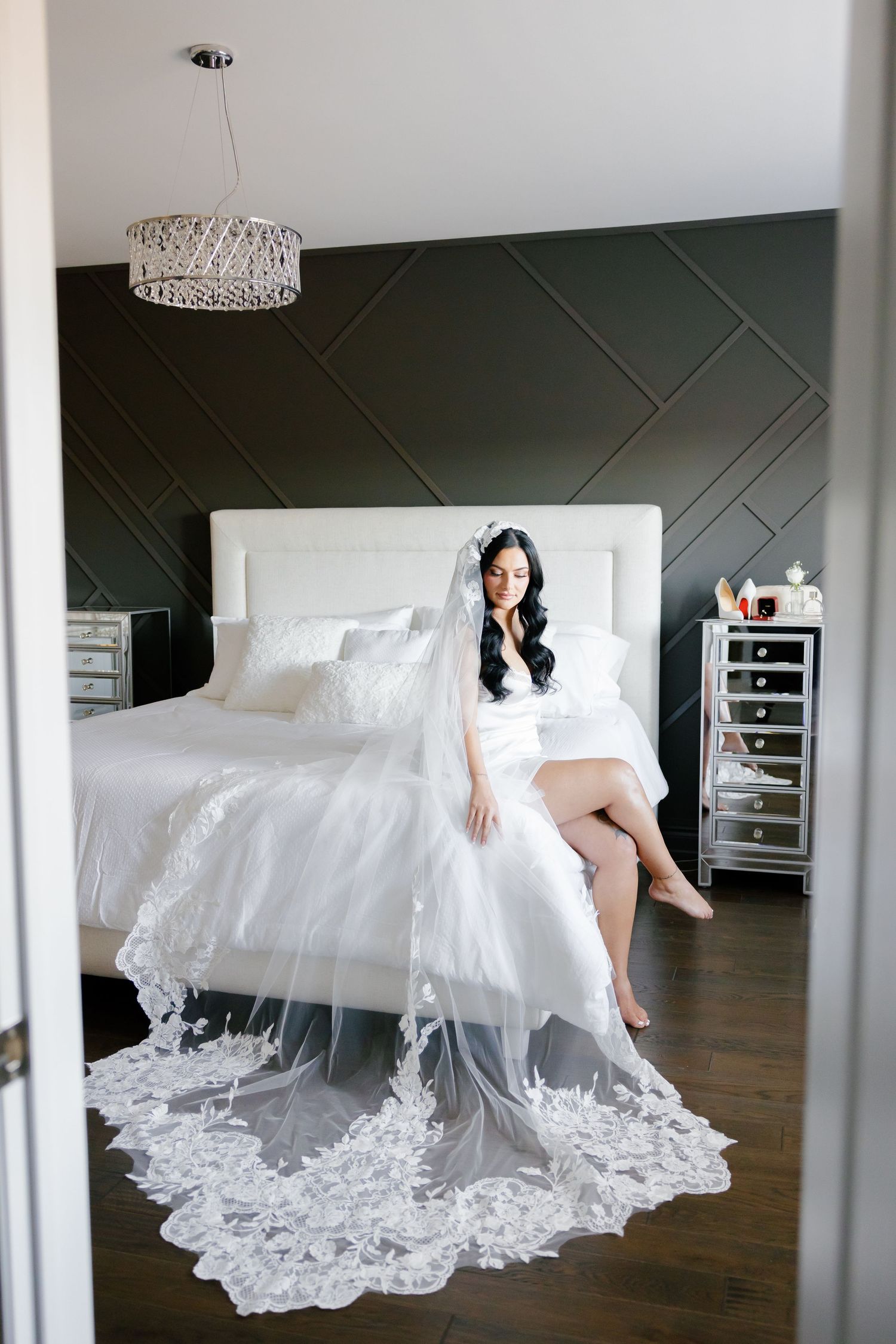 Modern bridal portrait on white bed with delicate lace veil against dark geometric wall.