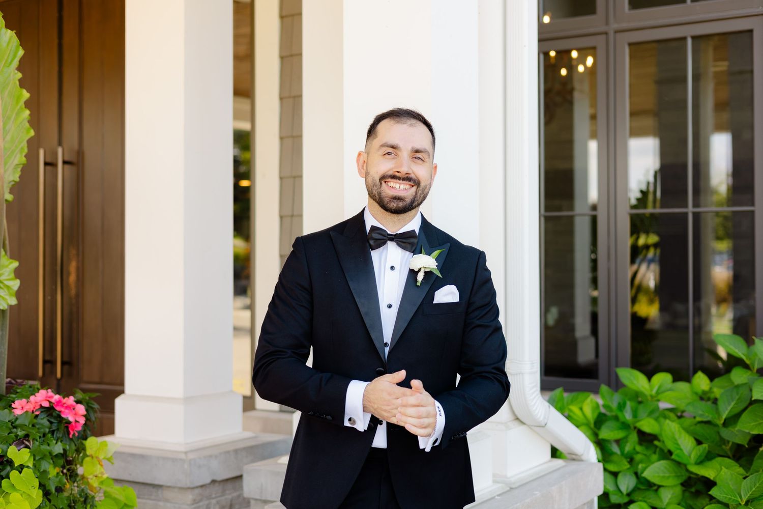 A well-dressed person in a formal black tuxedo poses on a porch with white columns and greenery.