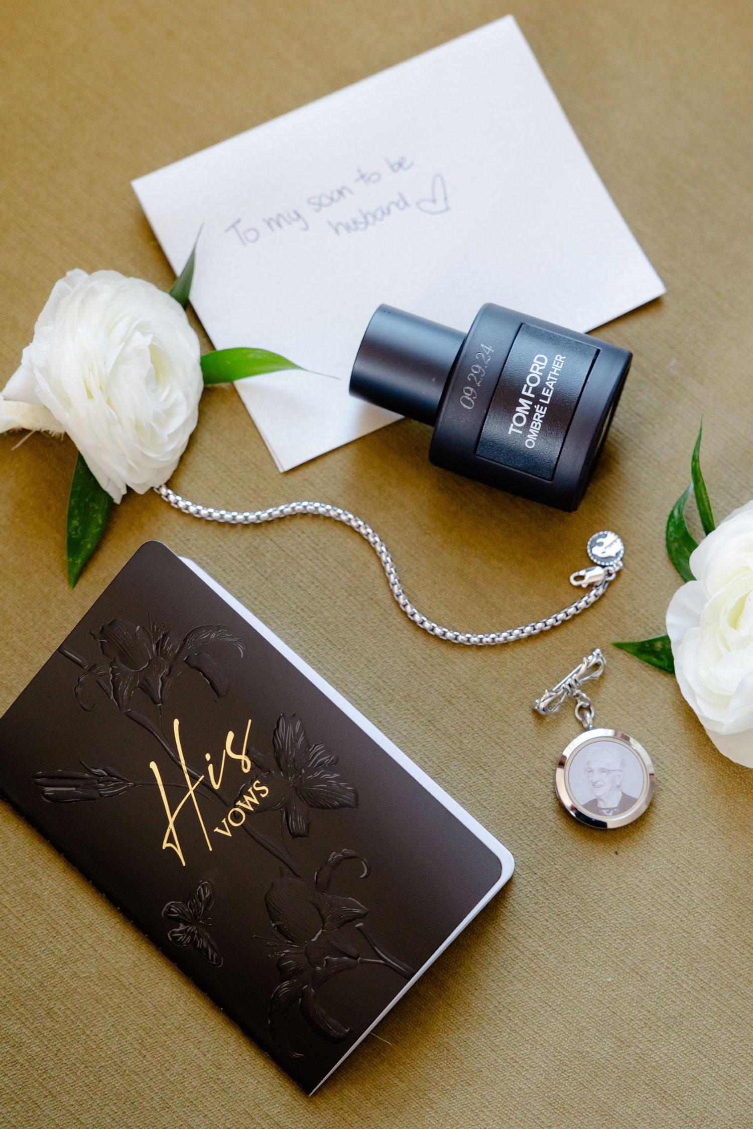 Wedding accessories including cologne, vows book, roses and jewelry arranged on brown background.