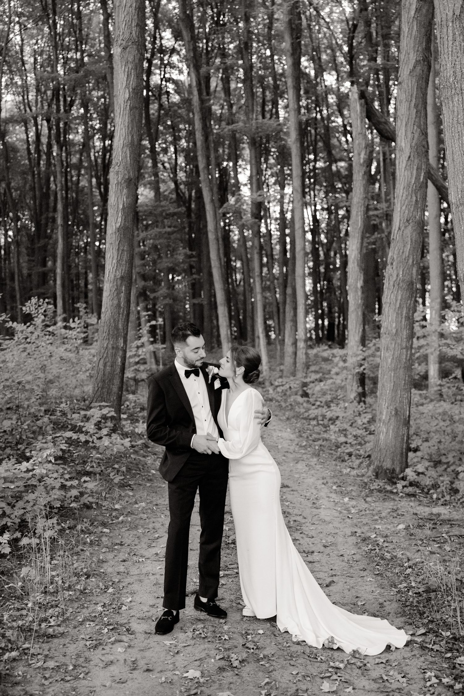 A black and white wedding portrait of a couple standing together on a forest trail.