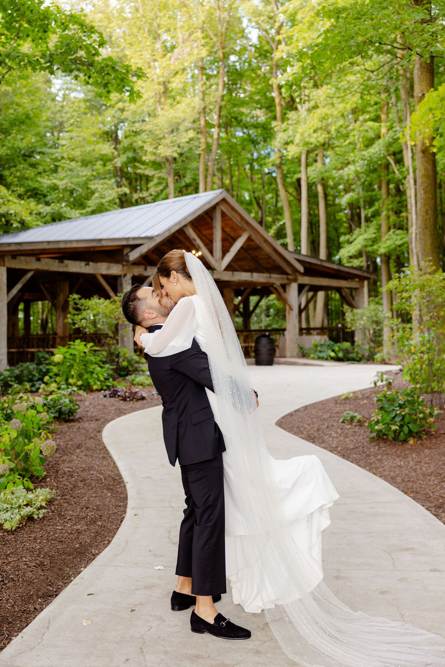 A joyful wedding couple dancing and embracing by a rustic pavilion in a wooded setting.