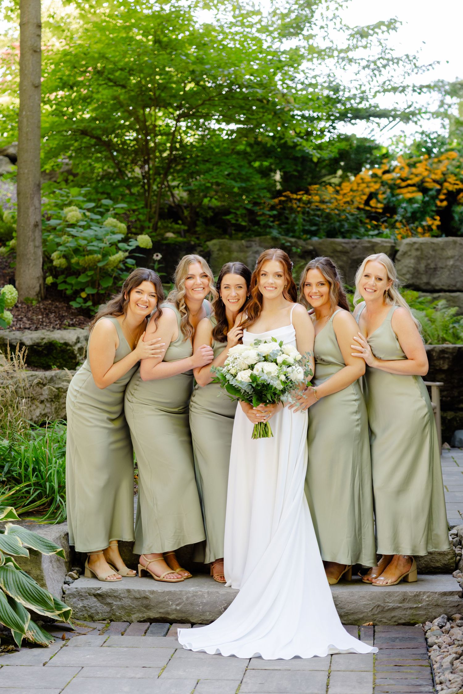 Wedding party stands on stone steps in garden setting with bride in white holding floral bouquet.
