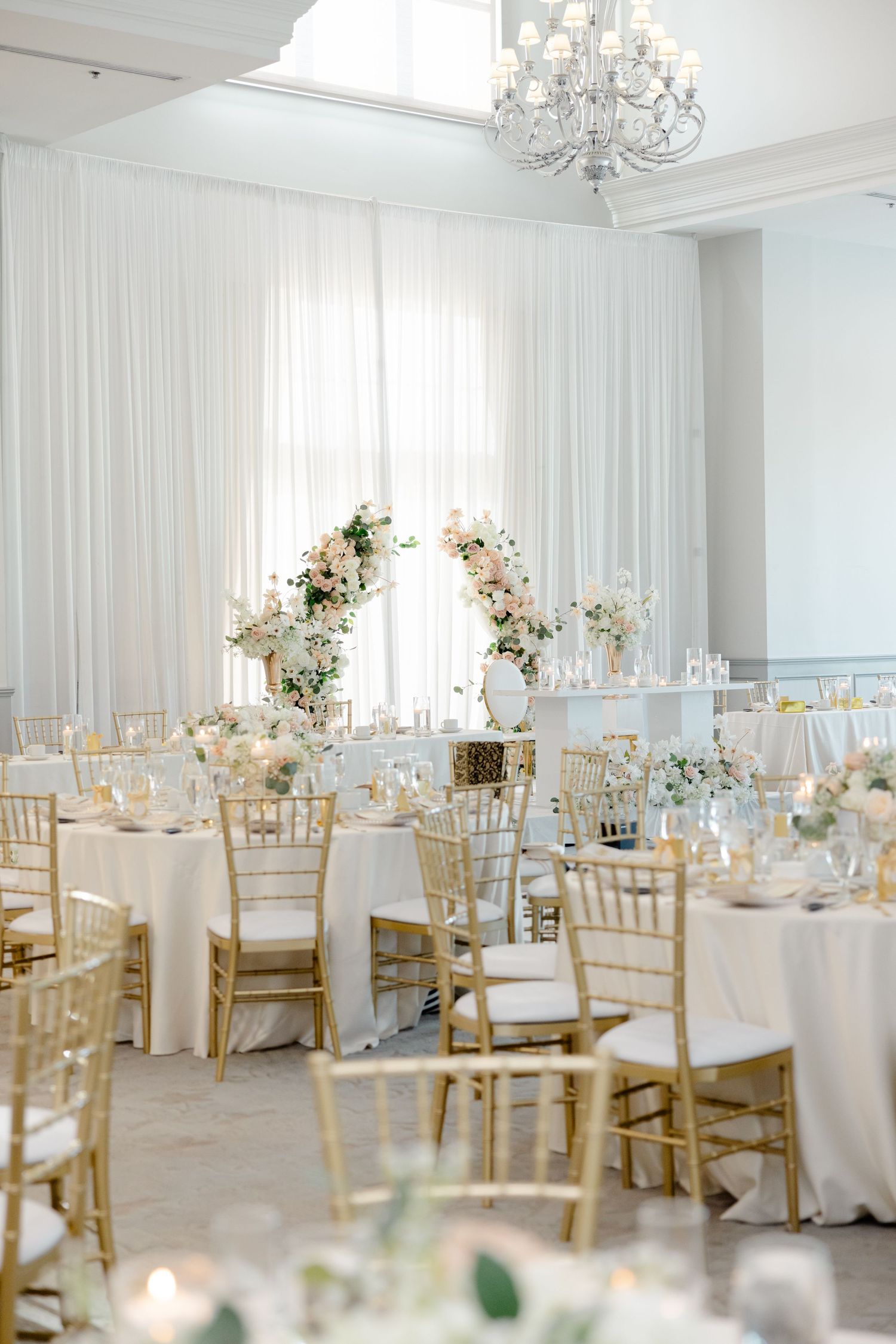An elegant wedding reception venue with white drapes, crystal chandelier, and gold chiavari chairs.