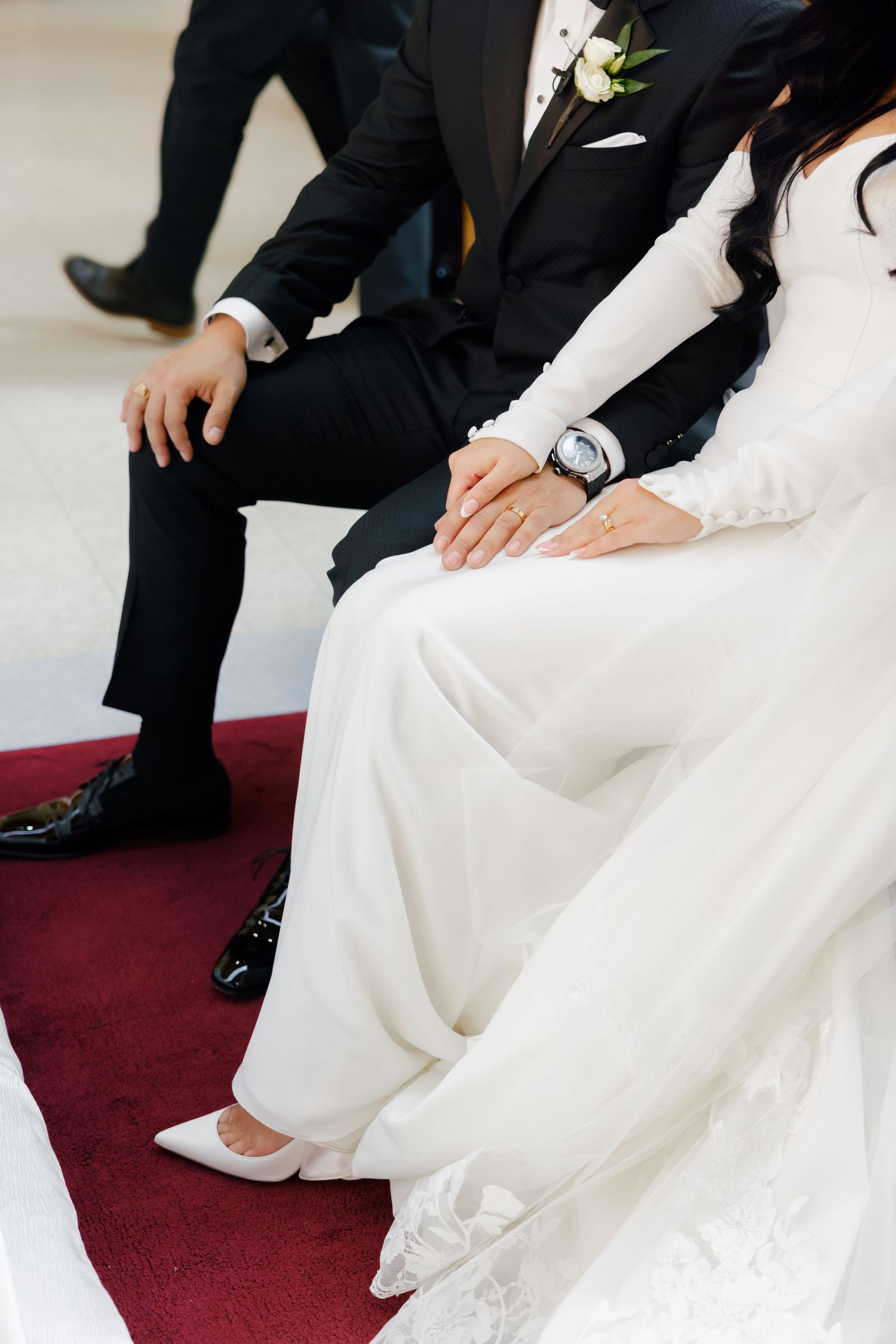 Close-up of wedding couple's formal attire and hands while seated on red carpet.