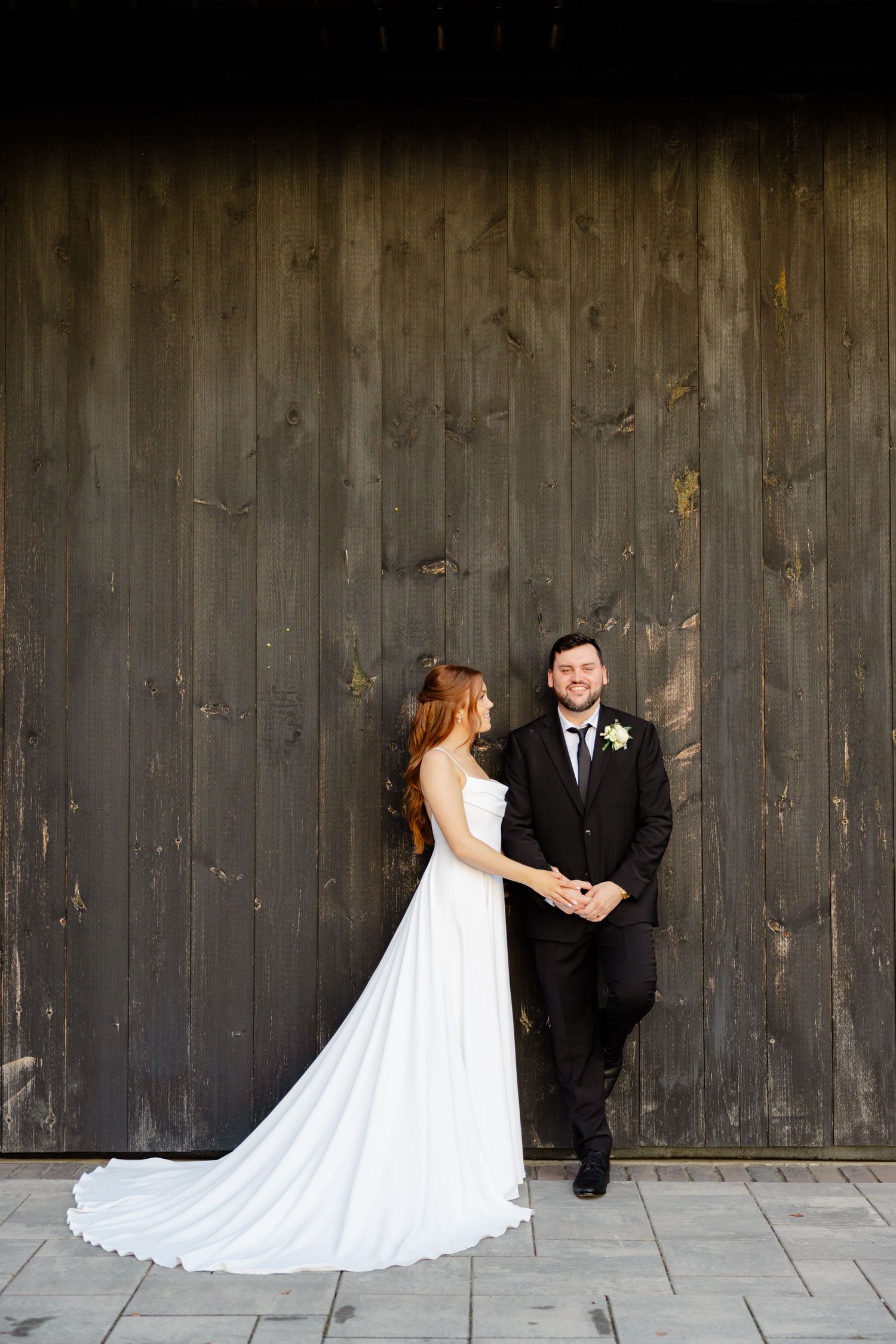 Wedding couple poses against rustic dark wooden wall with elegant white gown and classic tuxedo.