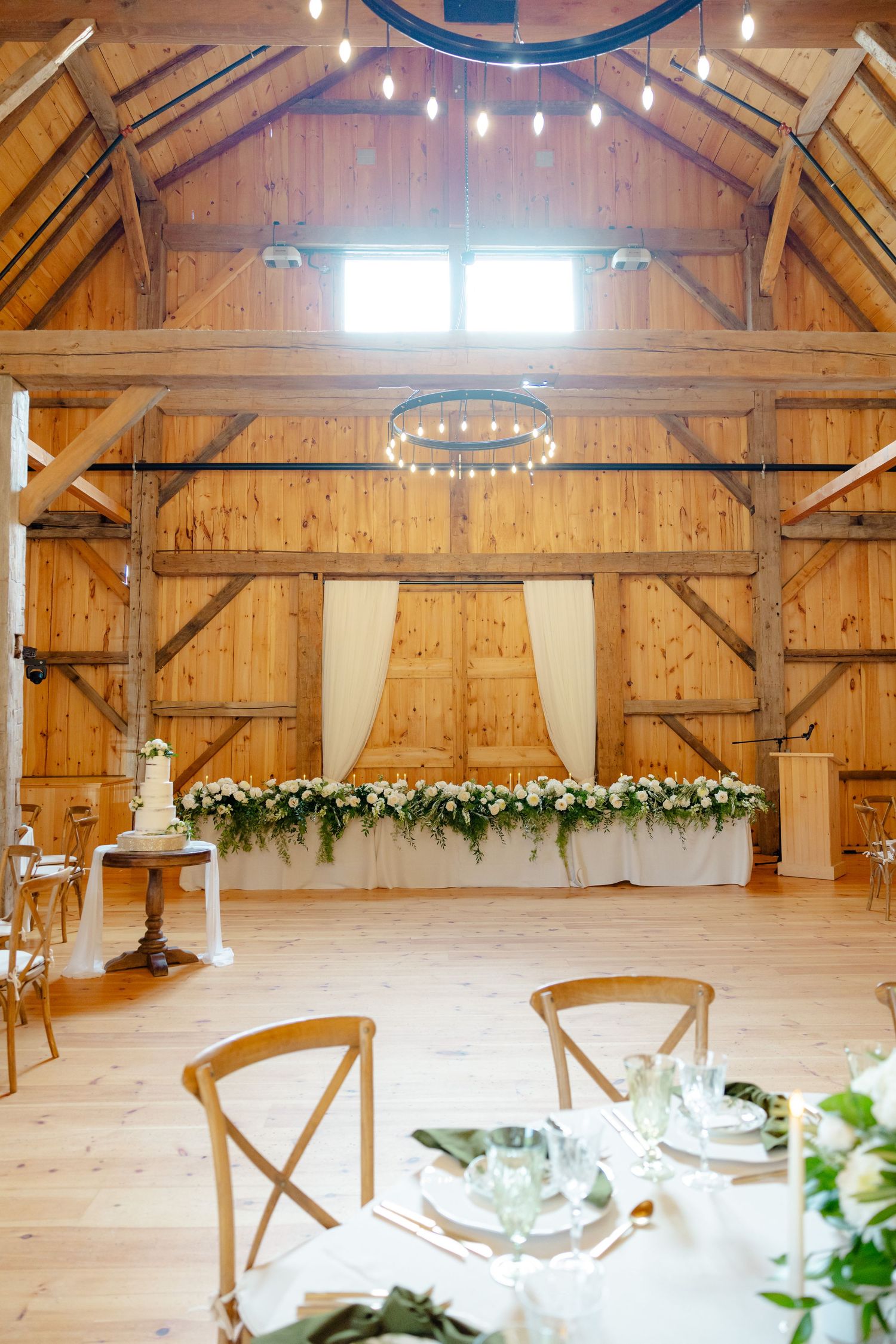 Rustic barn wedding venue interior with wooden beams, string lights and floral garland.
