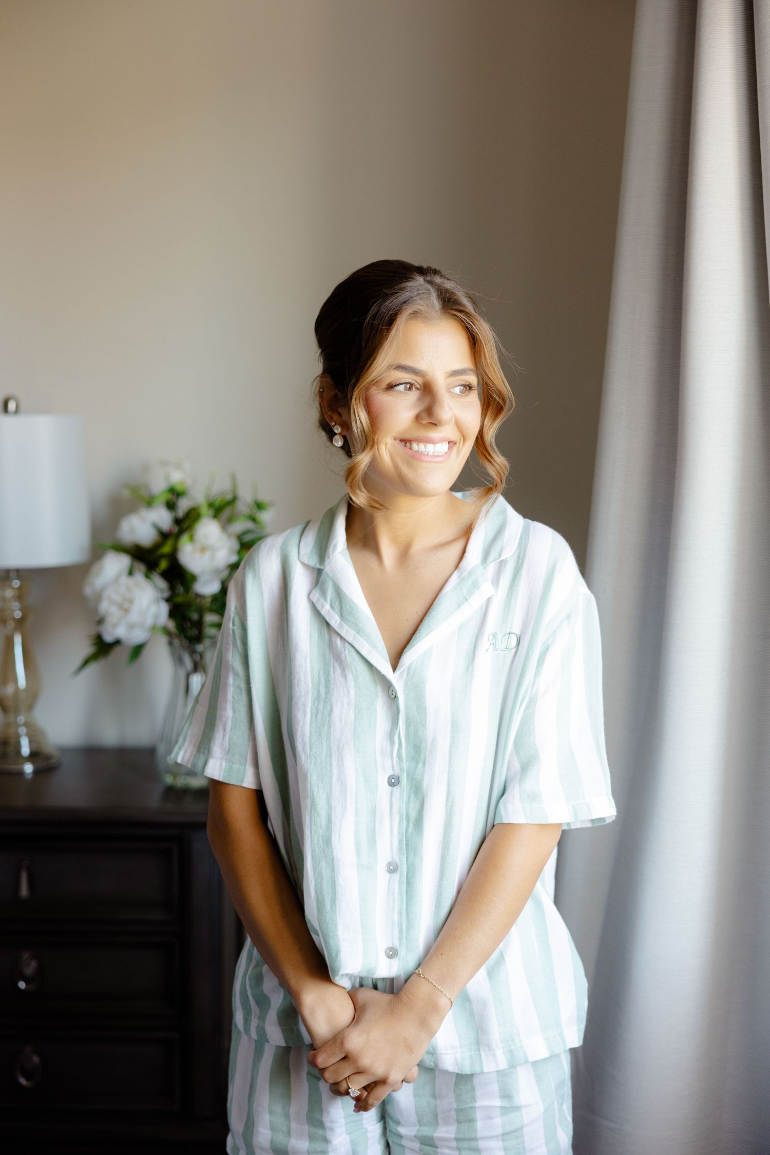 A person wearing a striped white pajama set smiles while standing in a bedroom near white flowers and a lamp.