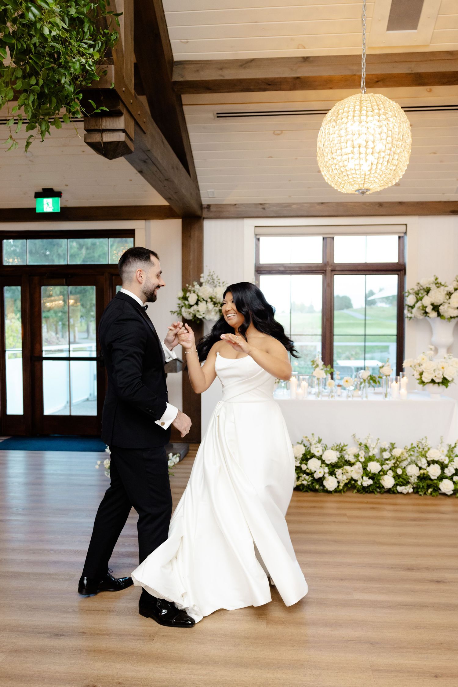 A couple dances together in a rustic venue with exposed beams and white floral decorations.