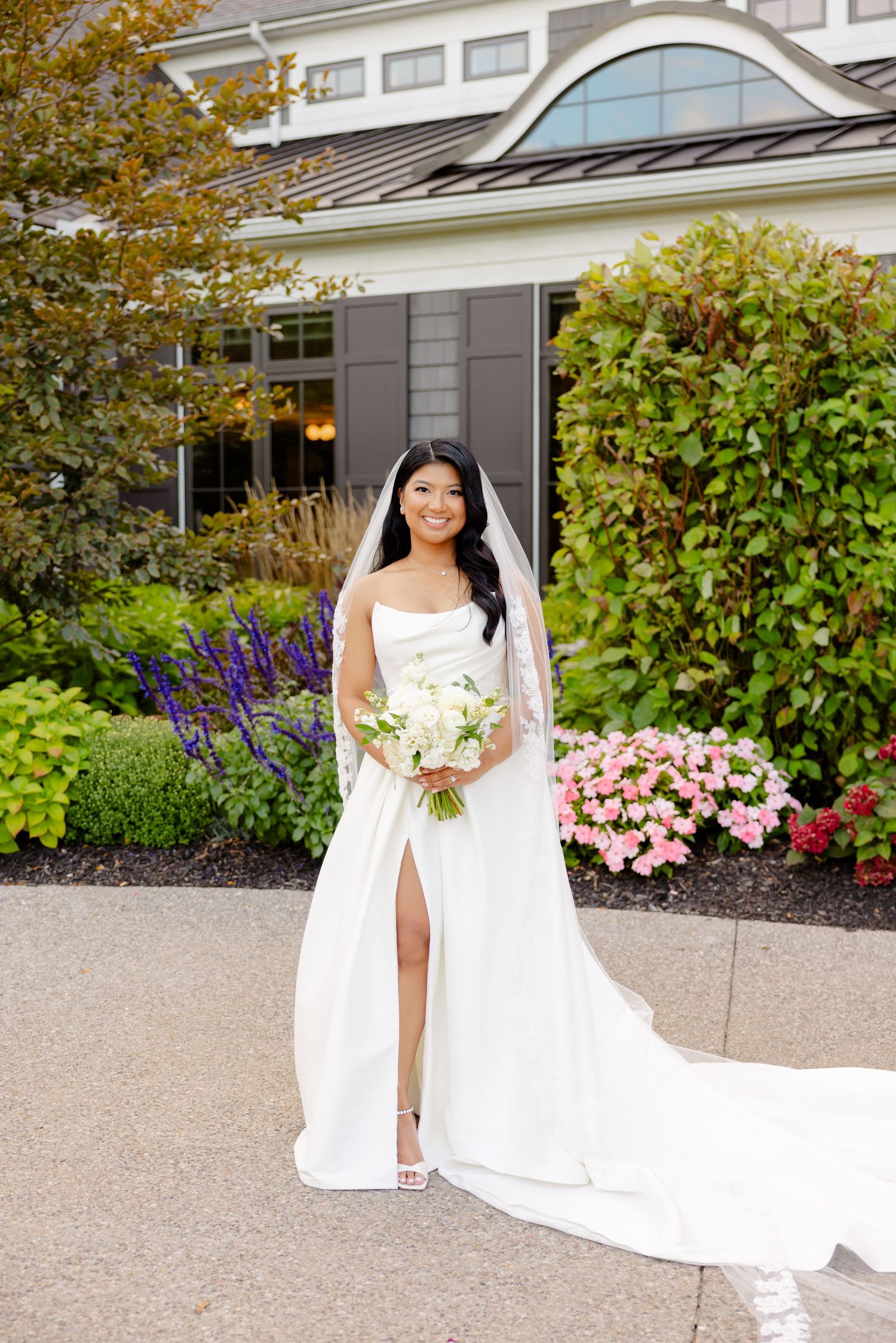 Modern bridal portrait featuring white wedding dress with thigh slit against grey house and colorful garden.