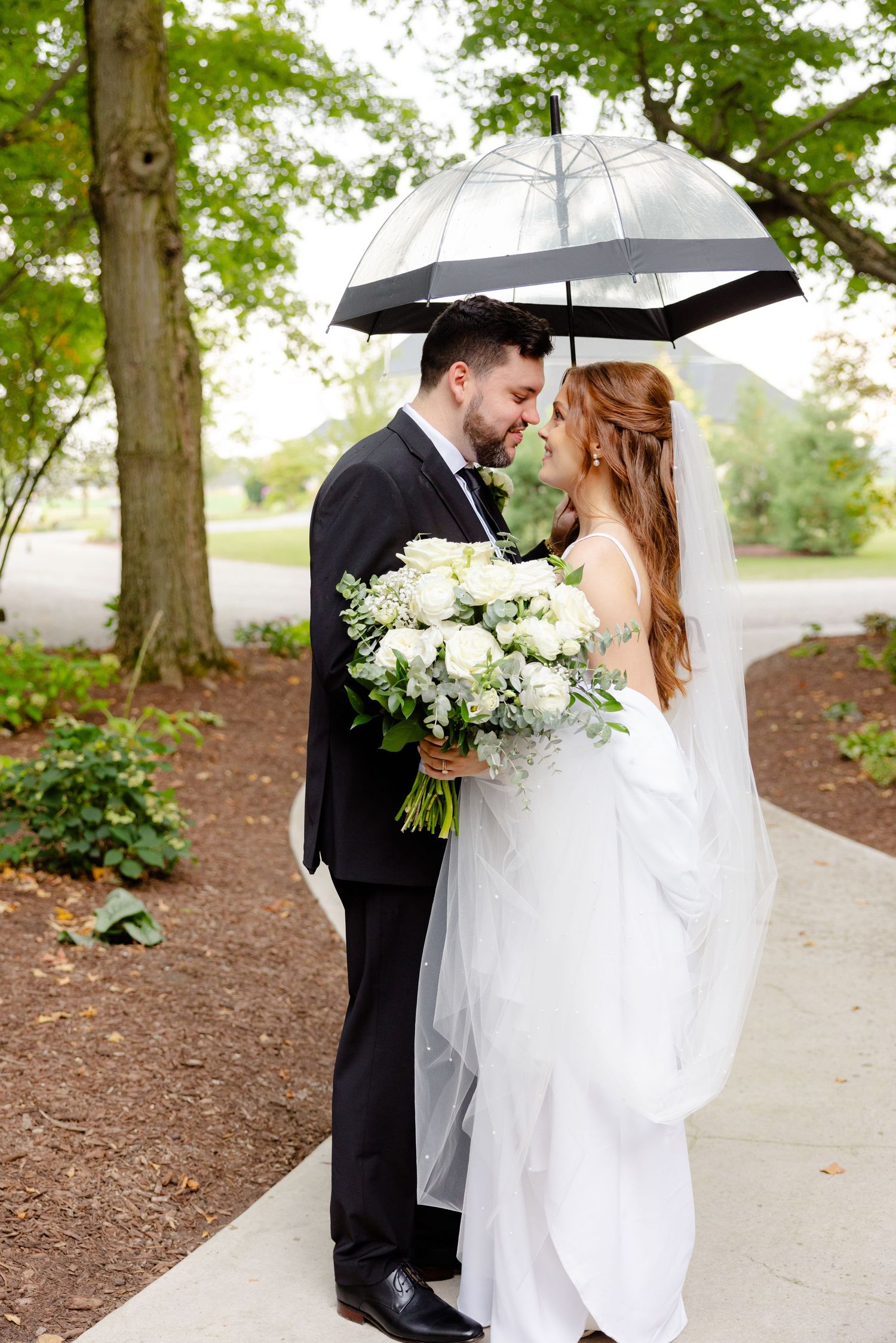 Wedding couple stands on a garden path under a clear umbrella with elegant white floral bouquet.
