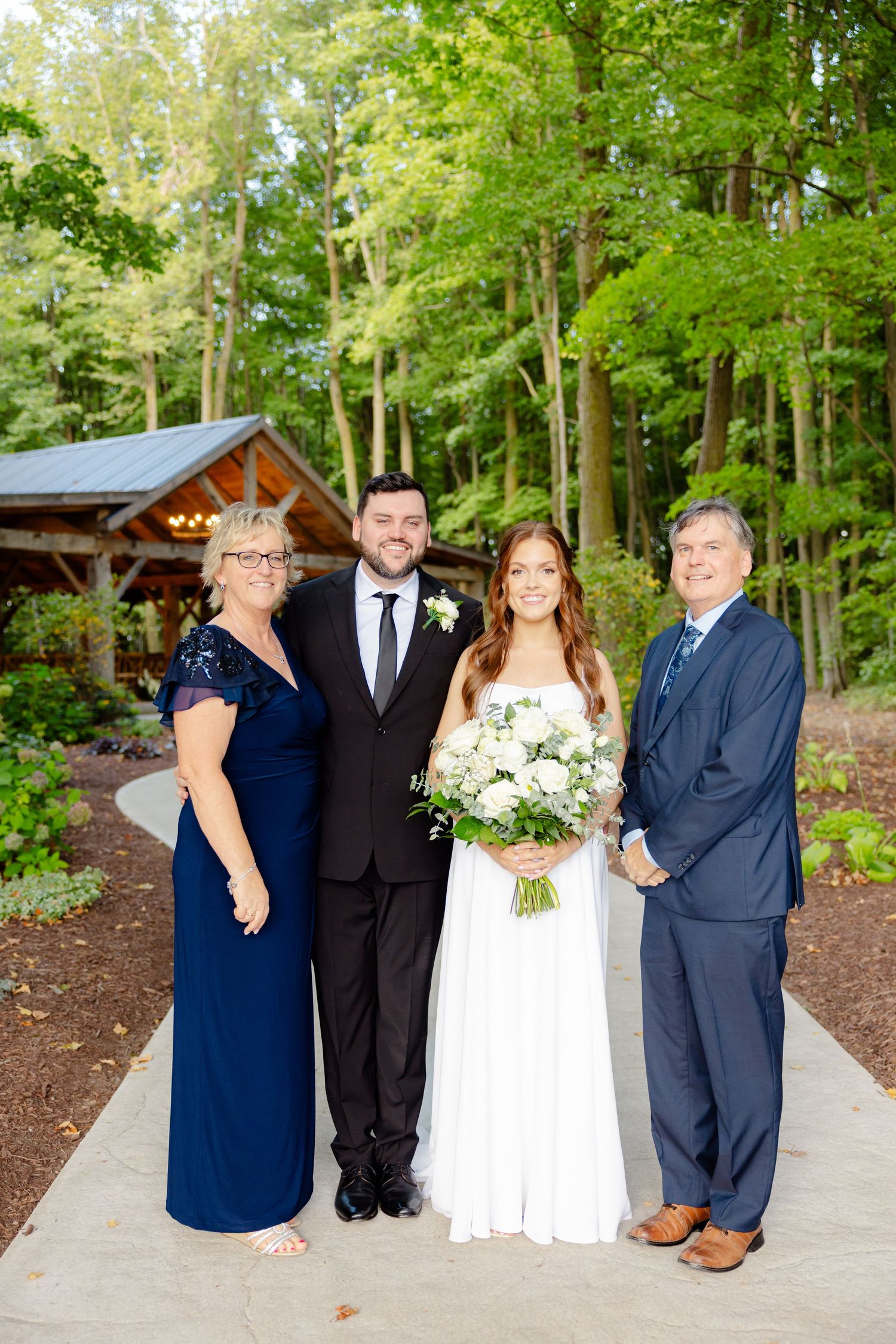 Family wedding portrait taken outside a rustic wooden pavilion surrounded by green forest.