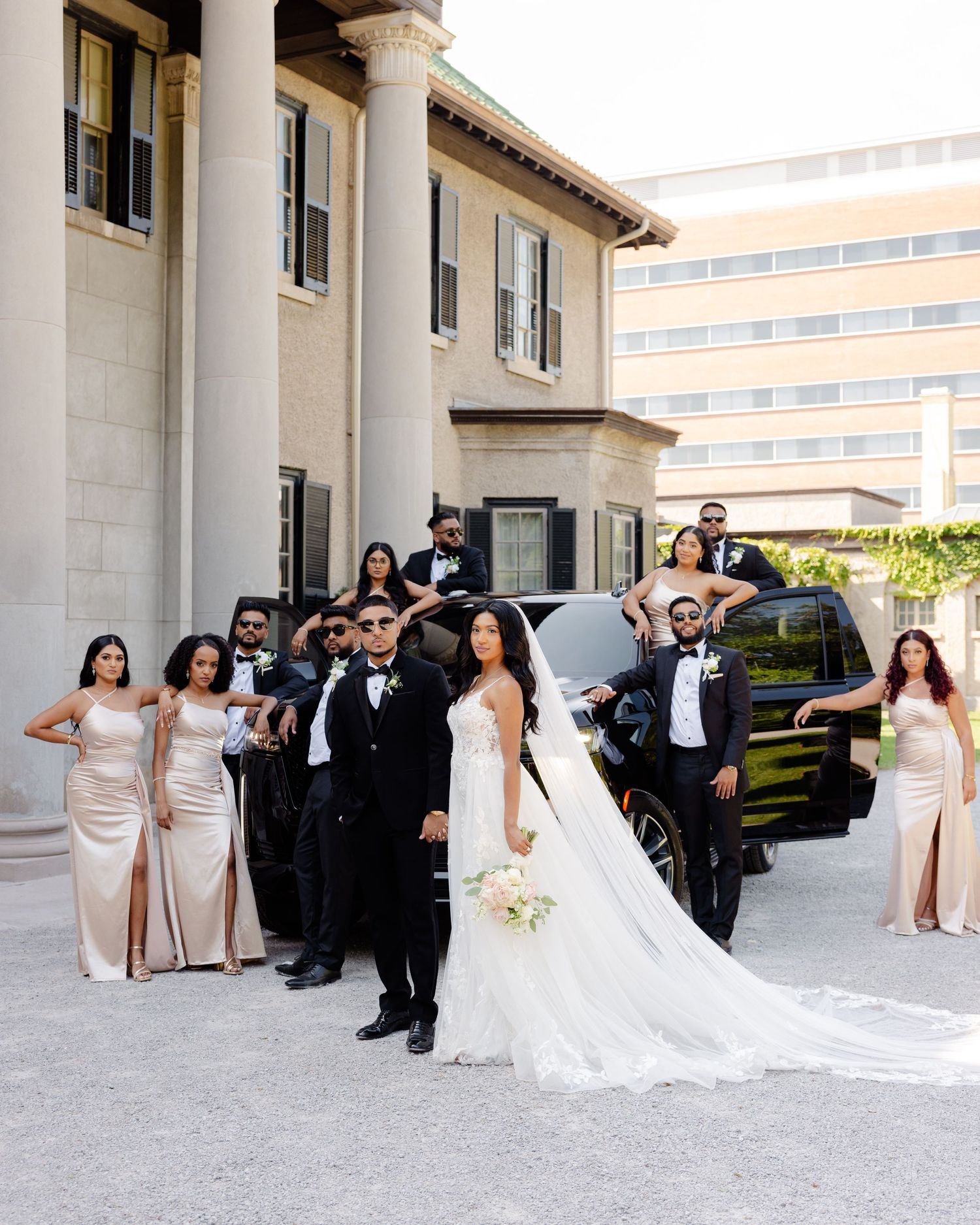 A wedding party in champagne and black formal attire poses outside a classic columned building.
