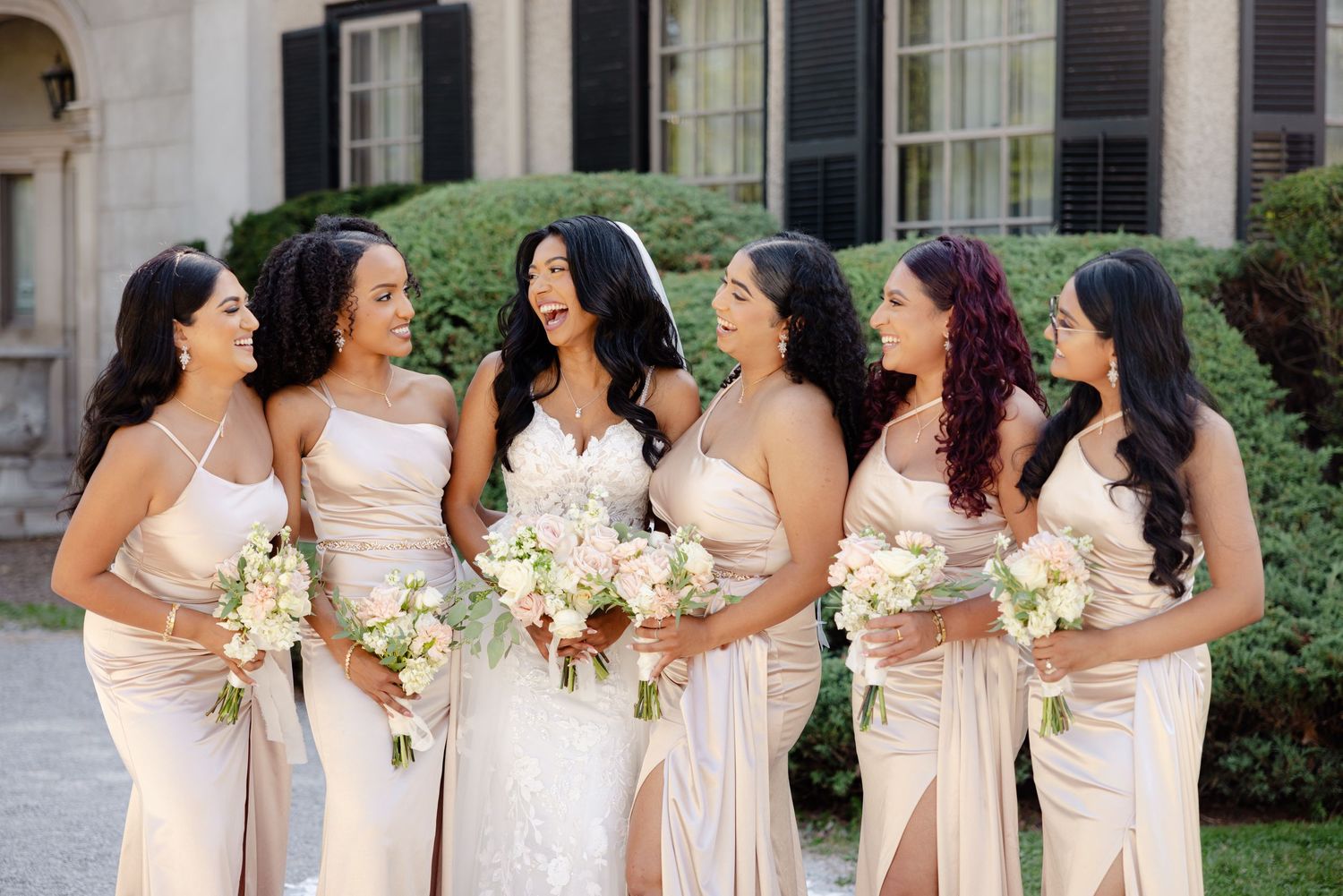 Bridal party shares candid moment in champagne gowns holding white floral bouquets.