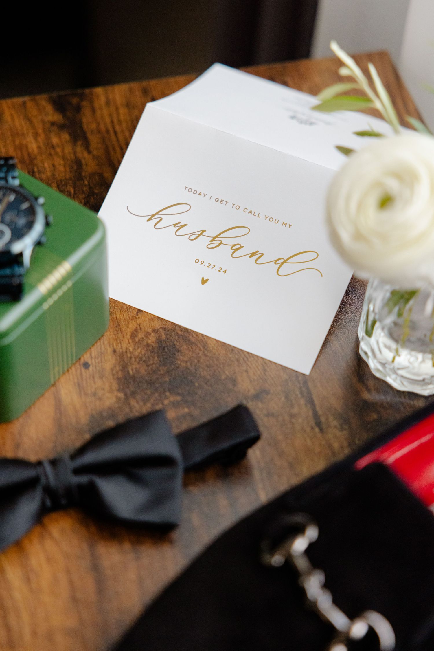 Wedding card with gold calligraphy next to black bow tie and green box.