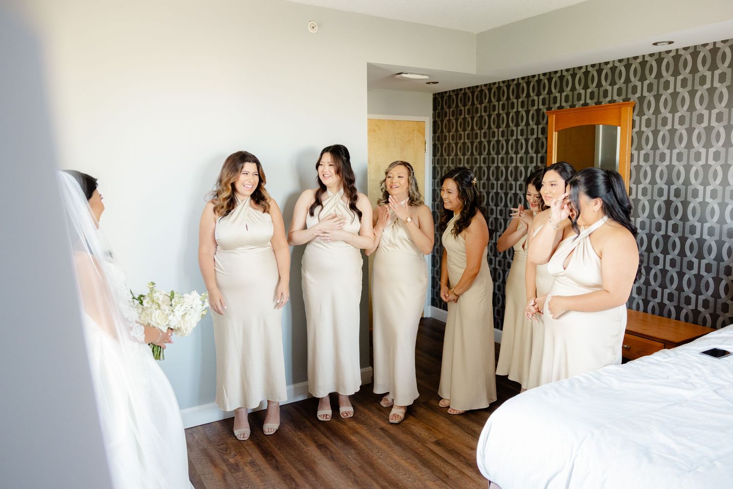 Bride in white gown with bridesmaids wearing cream dresses gather in hotel room for first look.