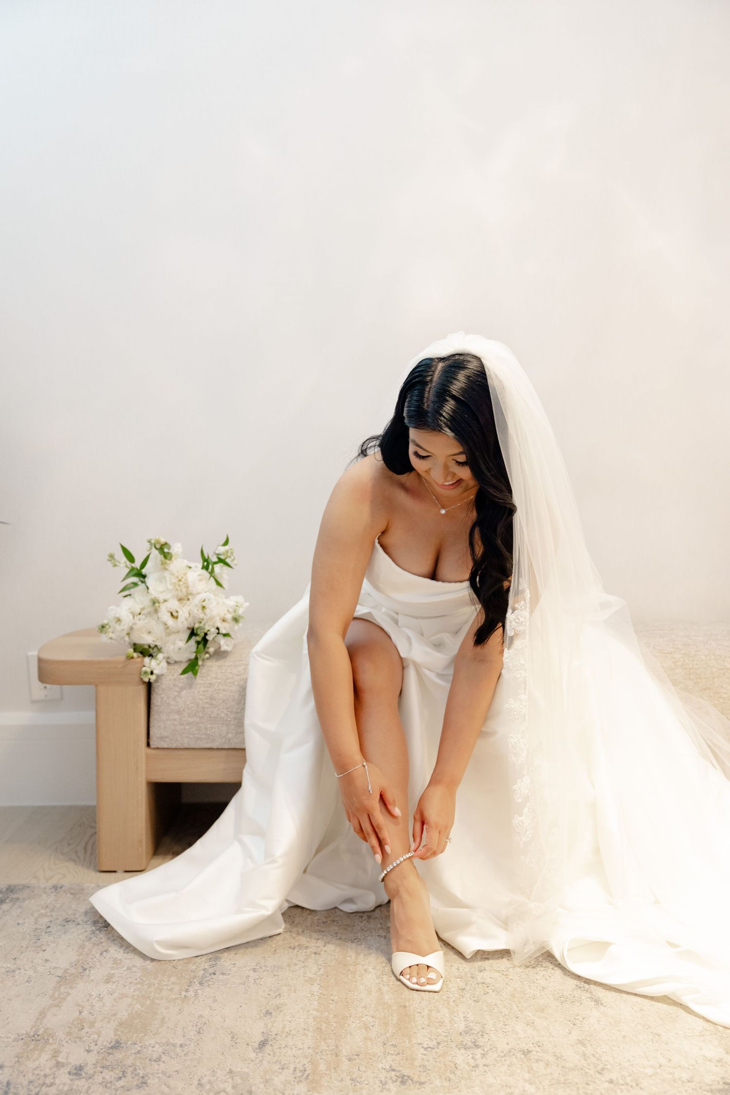 Bride in white dress adjusts her shoe while sitting in elegant natural light setting.