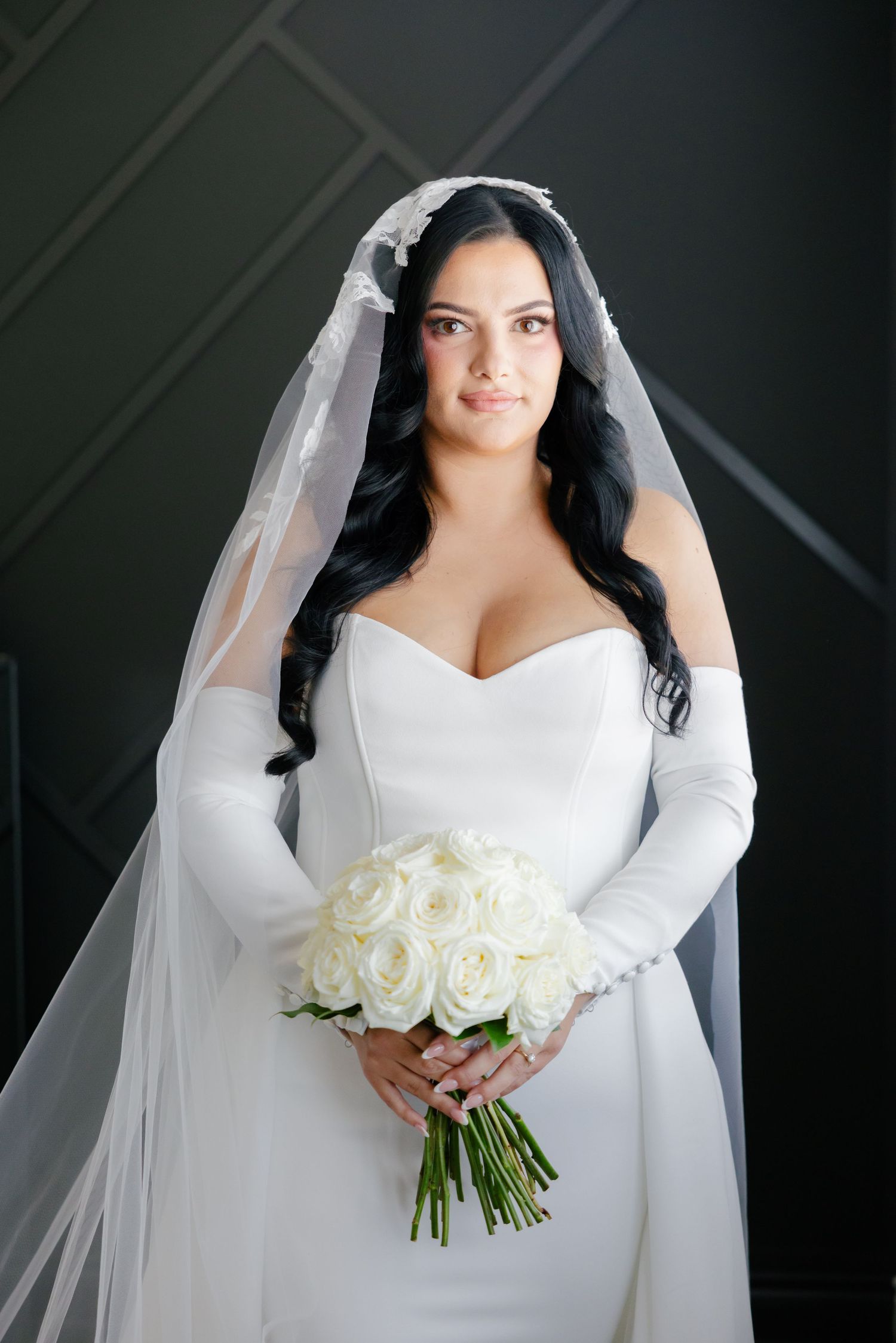 Classic bridal portrait with white roses and cathedral veil against geometric wall.