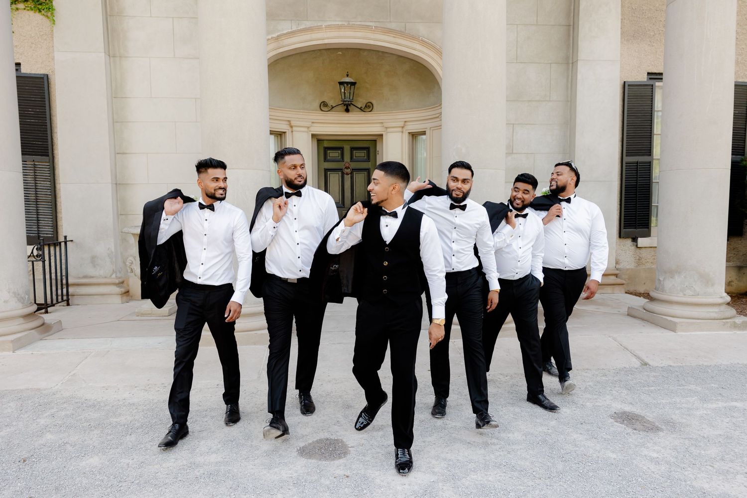 Wedding party of groomsmen in black and white attire walking and posing energetically outside.