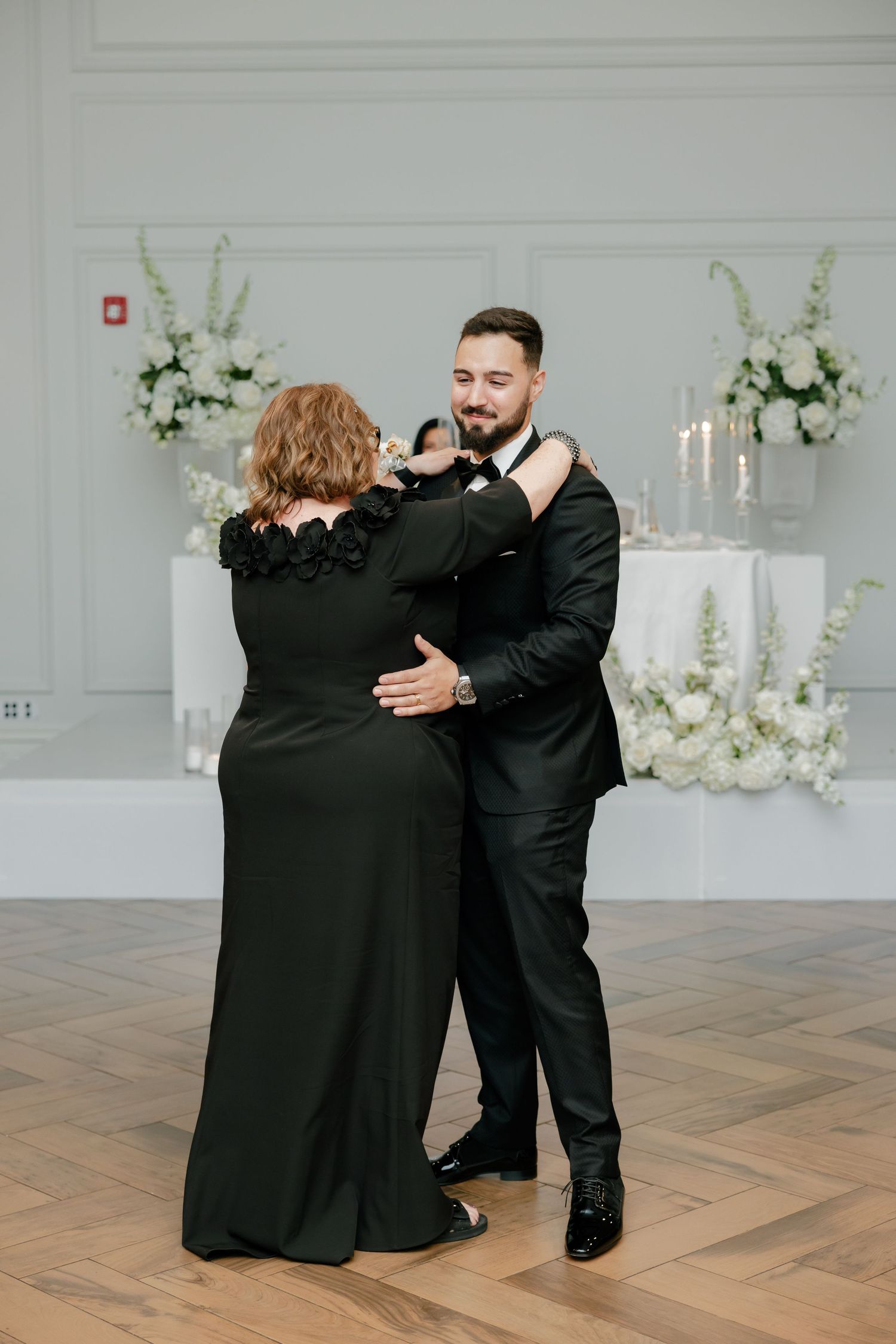 Two wedding guests share a dance in formal black attire on a classic herringbone wood floor.
