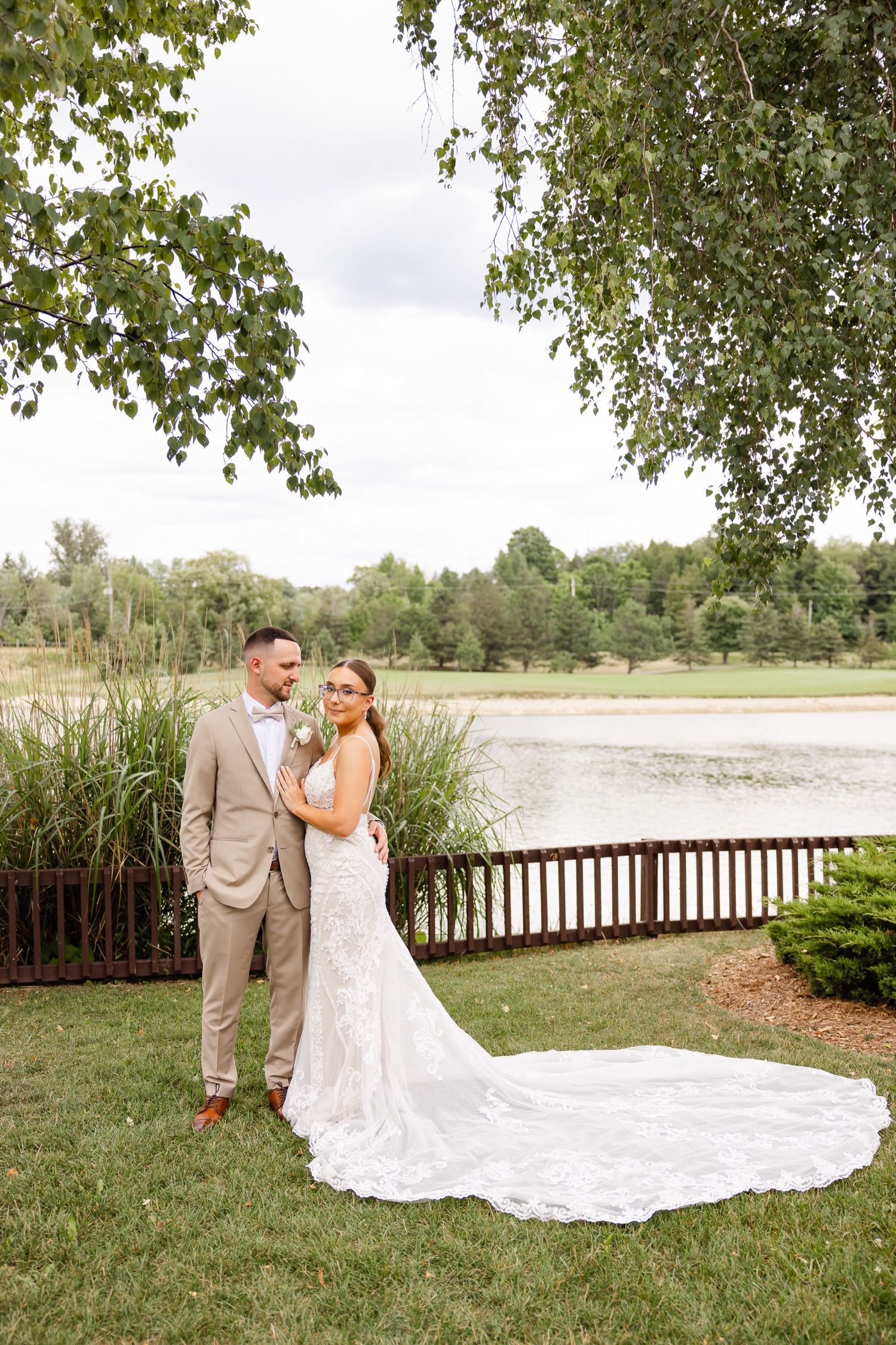 Elegant wedding portrait by a lake with bride's dramatic lace train extending across green grass.
