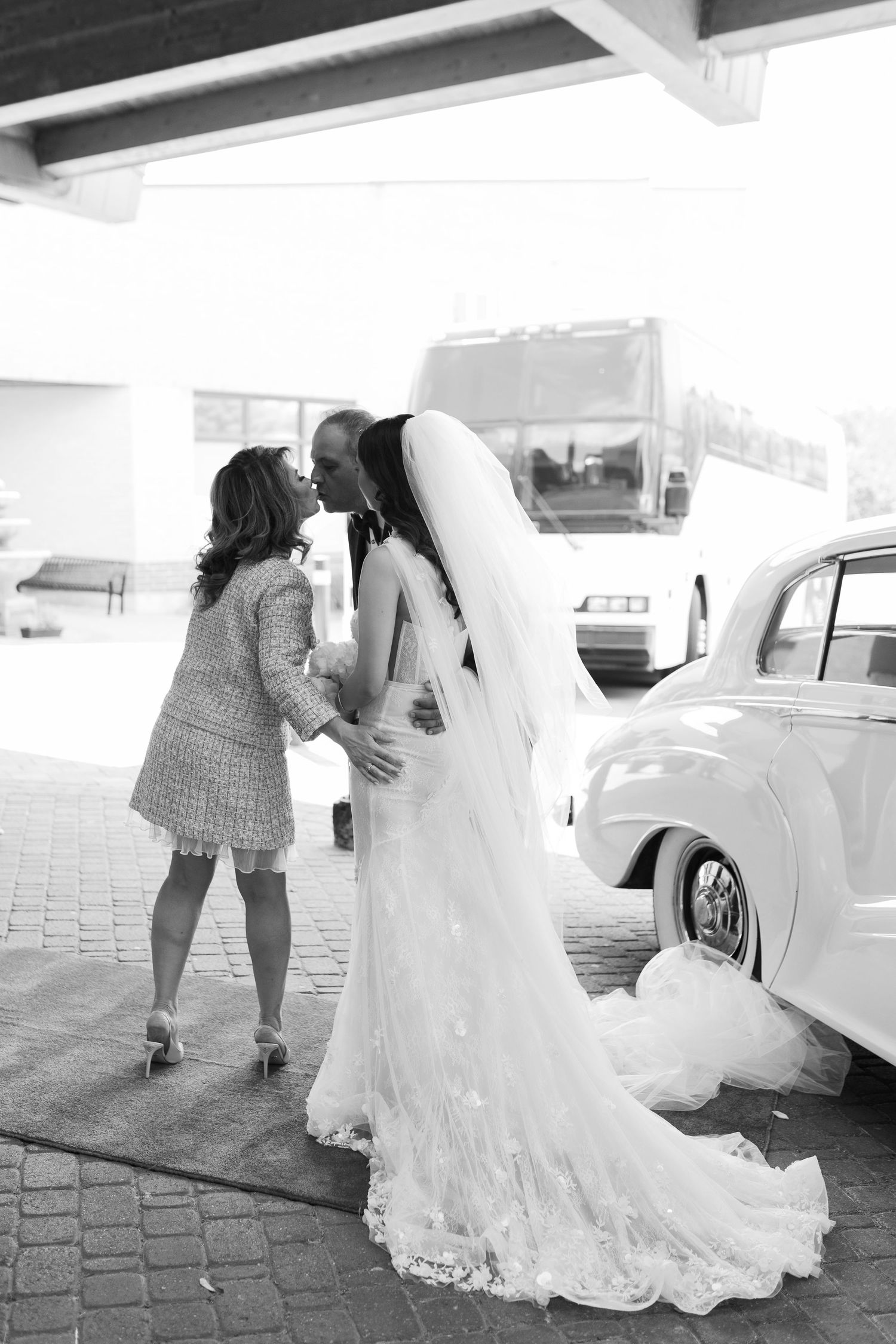 A tender moment between wedding guests by a vintage car in black and white photography.