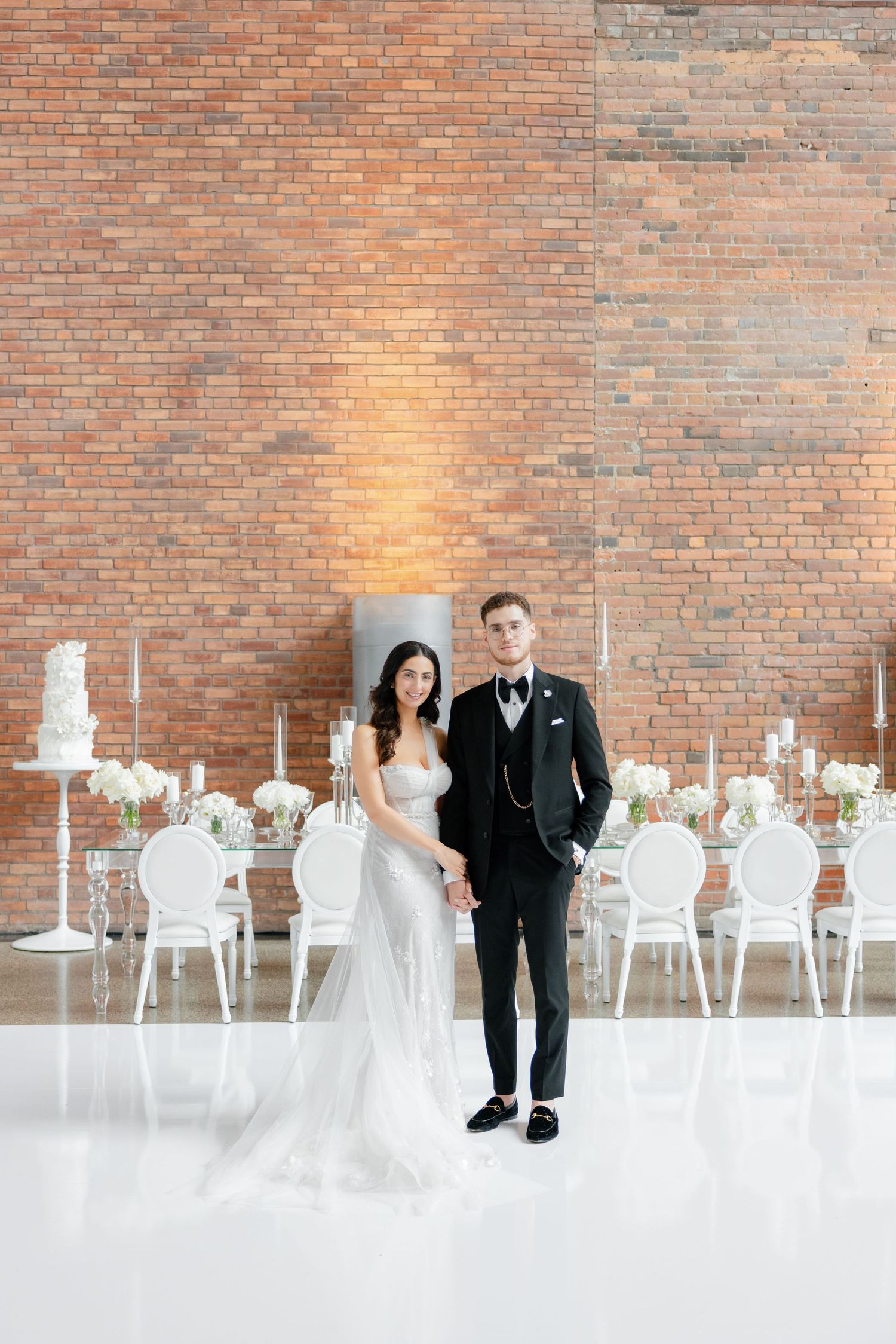 Couple standing in front of exposed brick wall with white wedding decor and candles.
