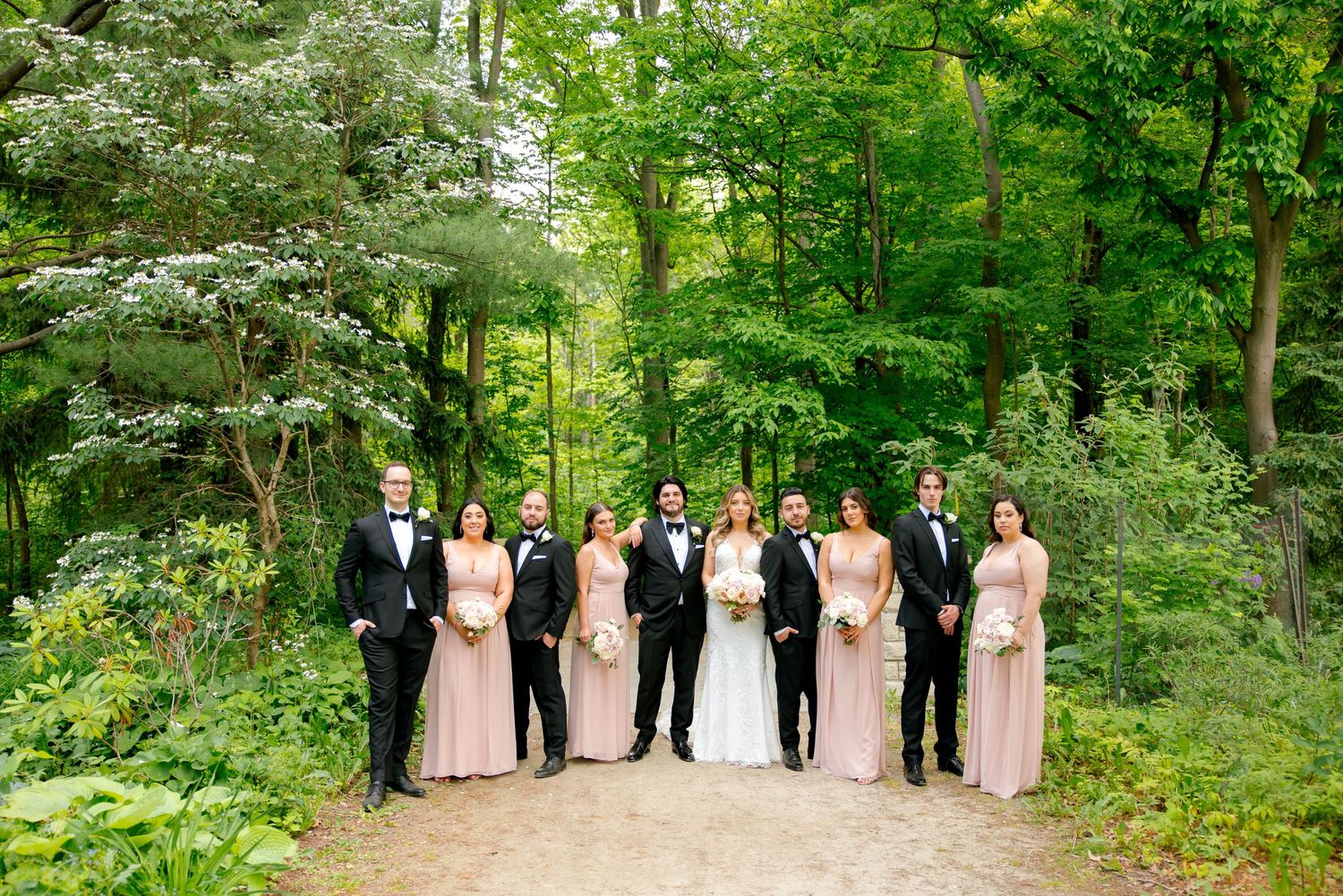 Full wedding party portrait on forest path with bridesmaids in pink and groomsmen in black tuxedos.