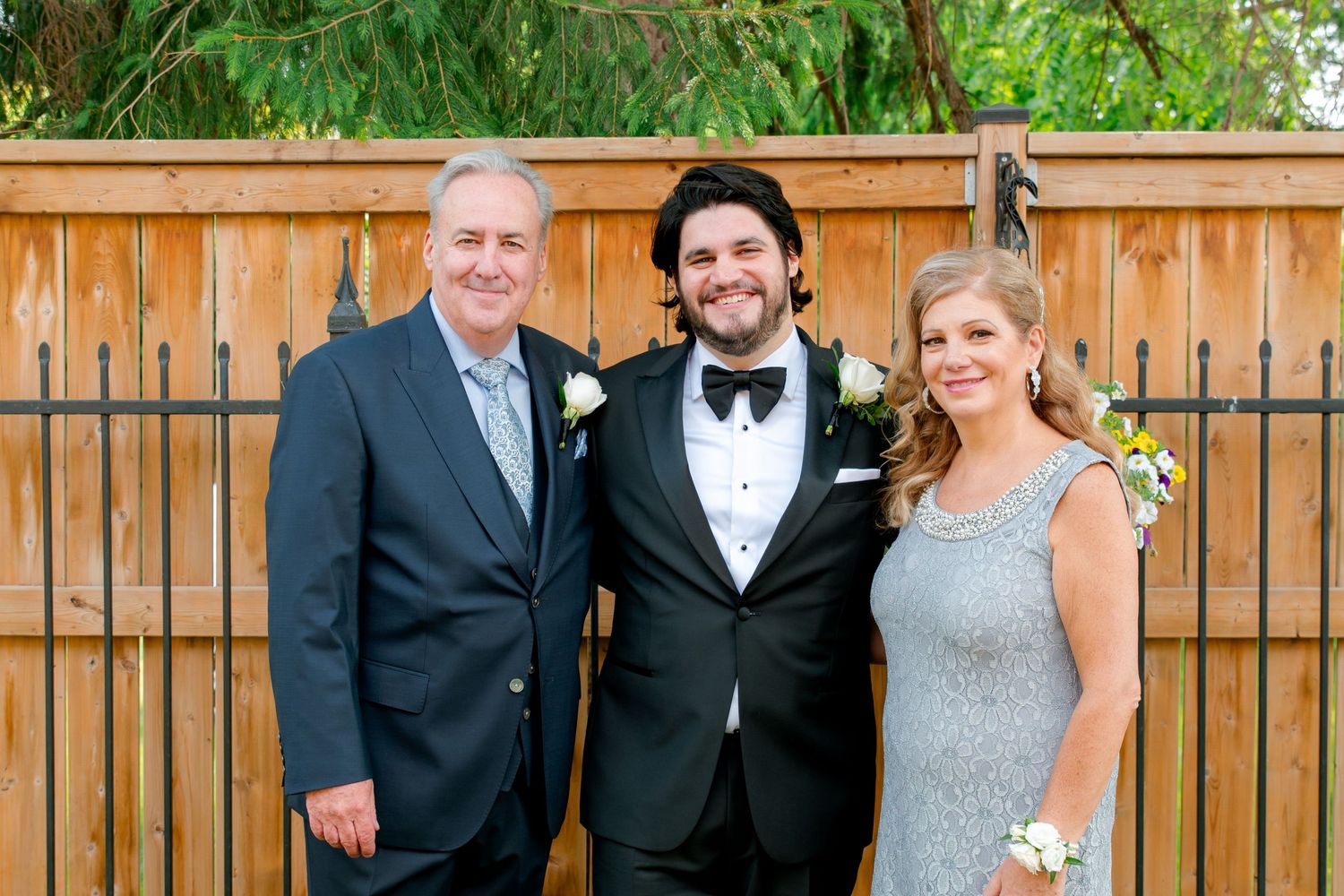 Elegant trio in formal attire pose together against wooden fence backdrop at outdoor gathering.