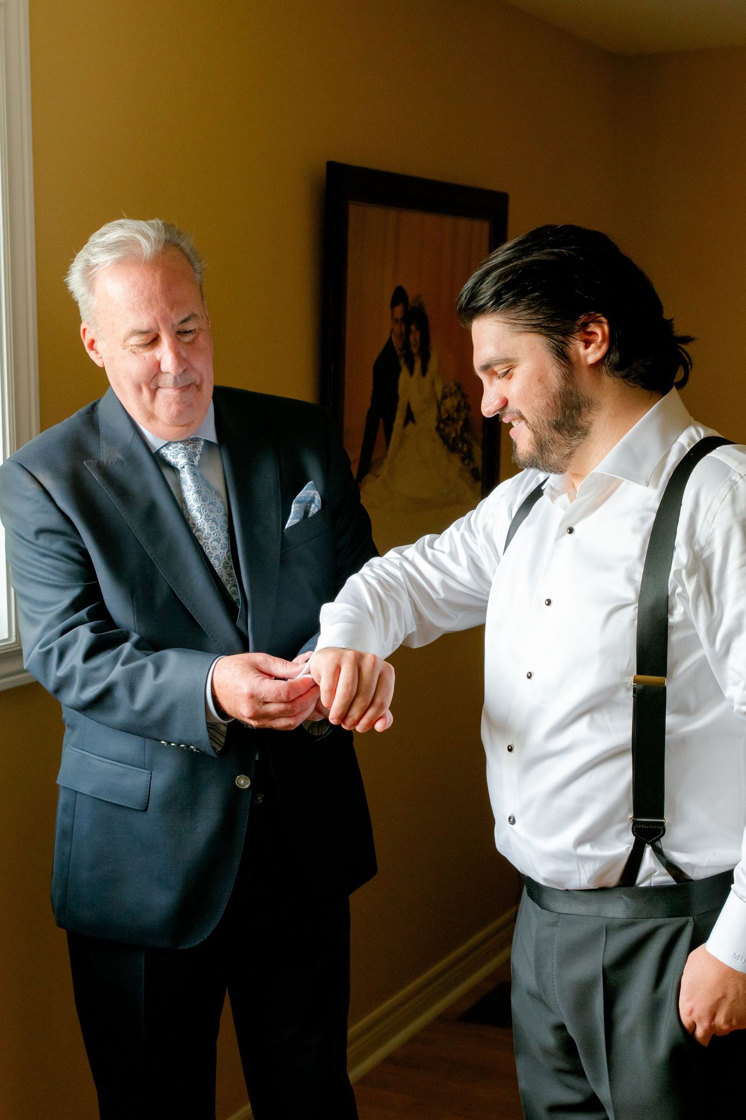 Two men getting ready before a formal event, one helping the other with cufflinks in a warmly lit room.