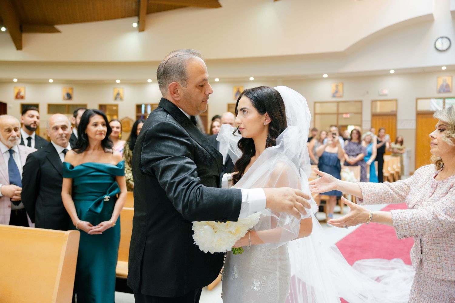Tender moment during a wedding ceremony as the bride's veil is adjusted in a church setting.