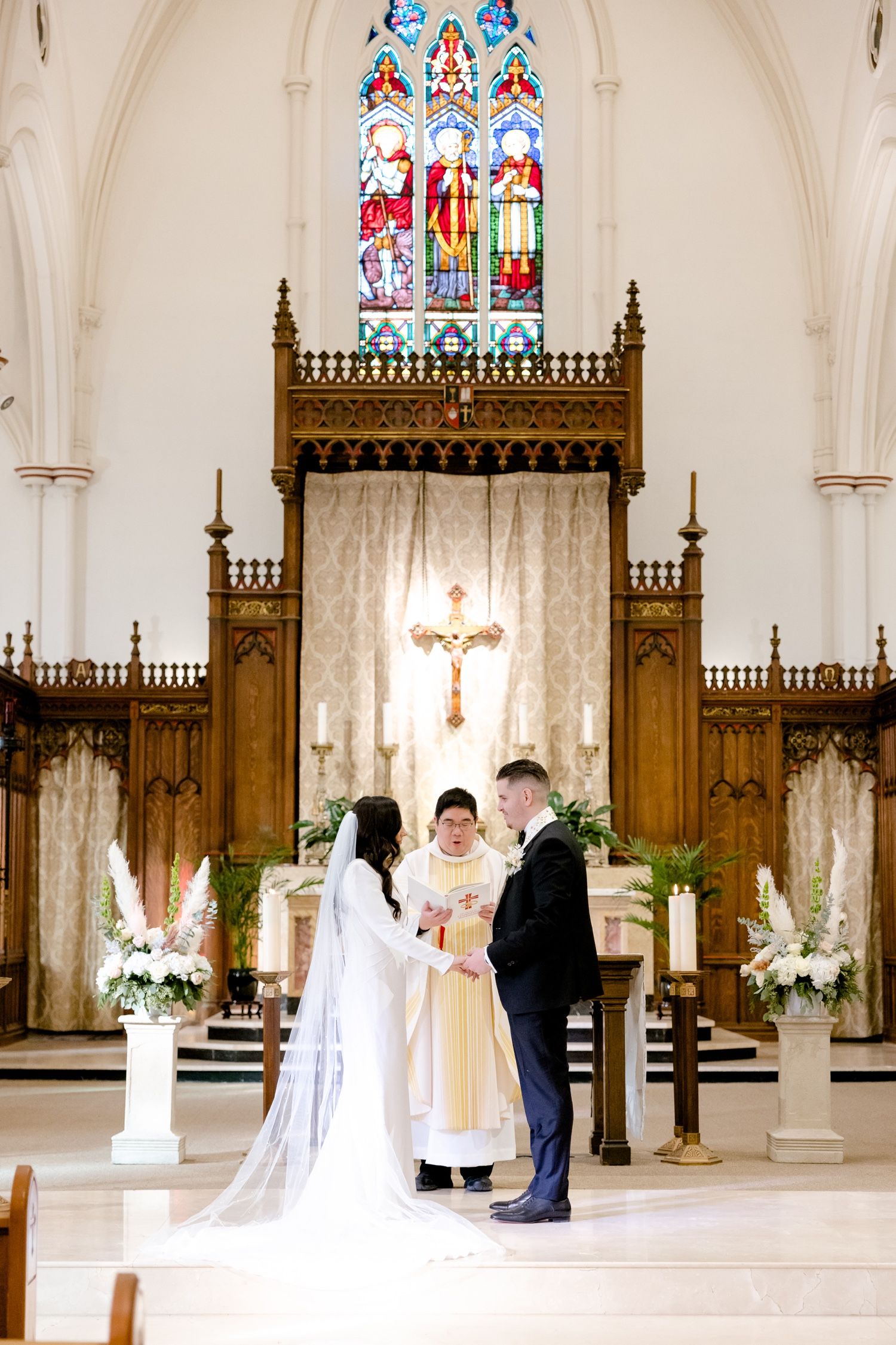A wedding ceremony takes place beneath ornate wooden church architecture and colorful stained glass windows.