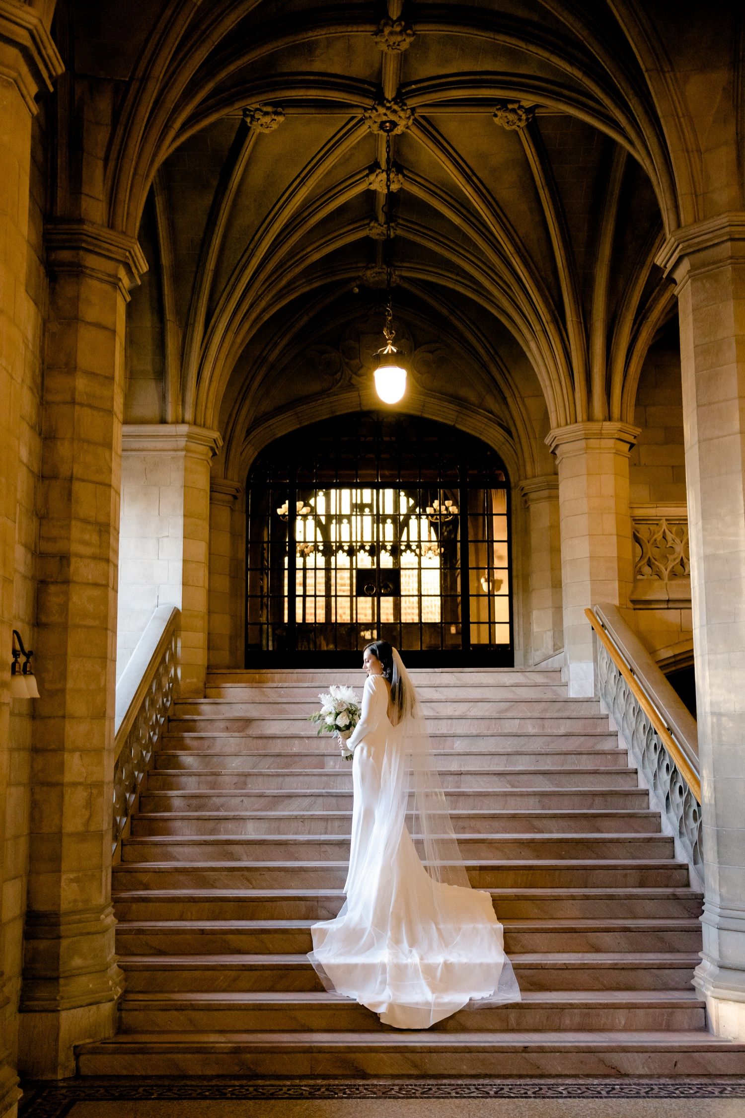 A bride in a flowing white gown stands on grand Gothic stone staircase with vaulted ceilings.