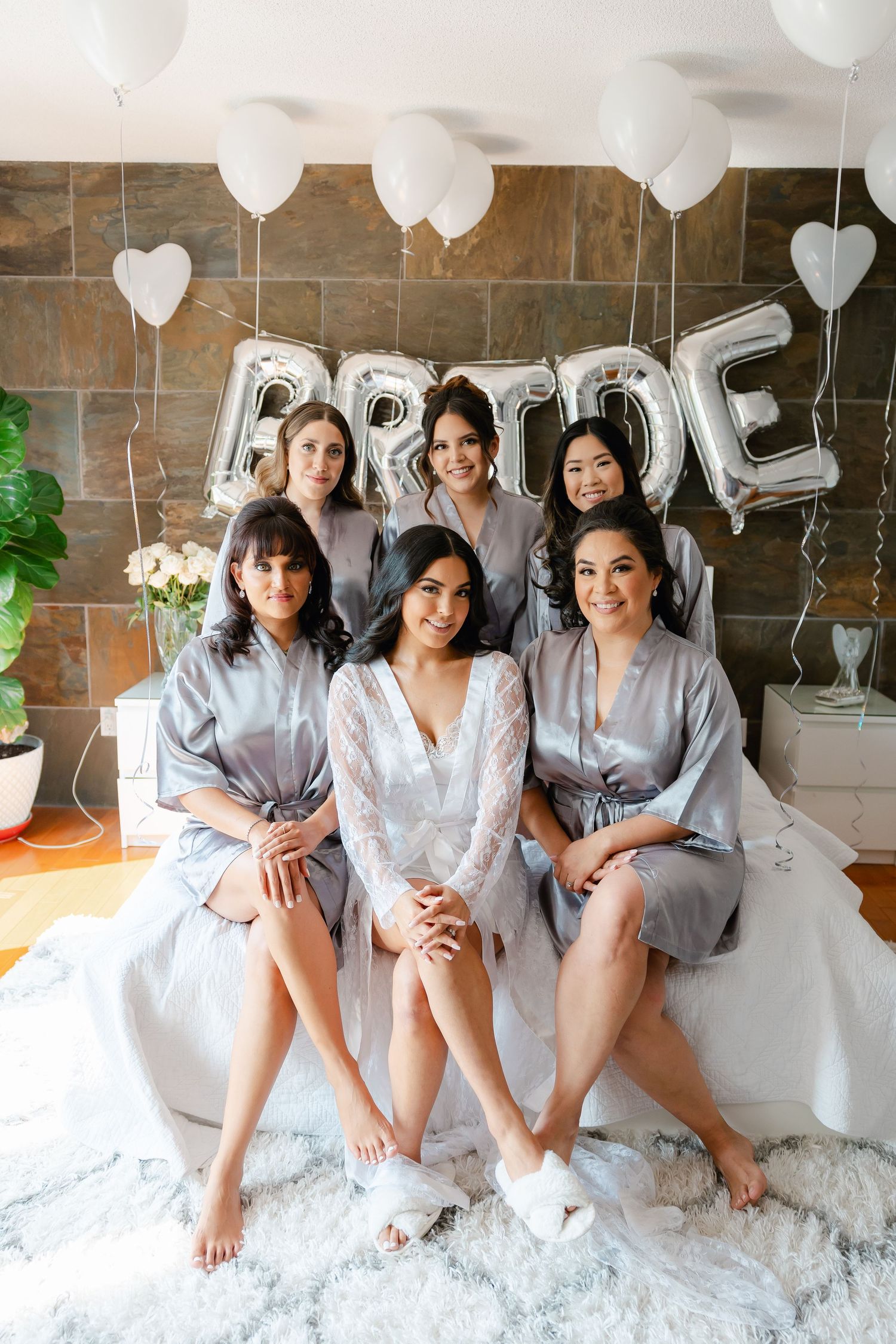 A bridal party in matching gray robes poses with silver balloon letters and white decorations.