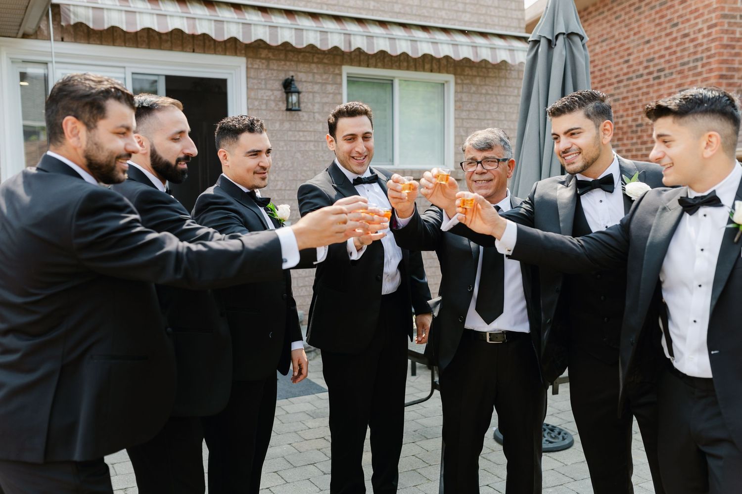 Groomsmen in formal attire raise glasses for a celebratory toast on brick patio.