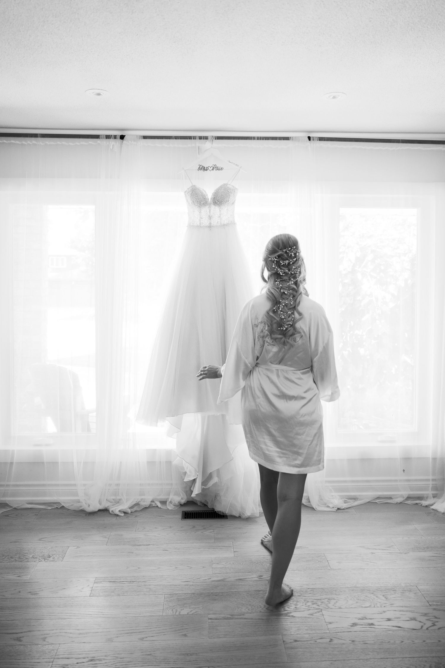 Black and white photo showing wedding dress hanging in window light.