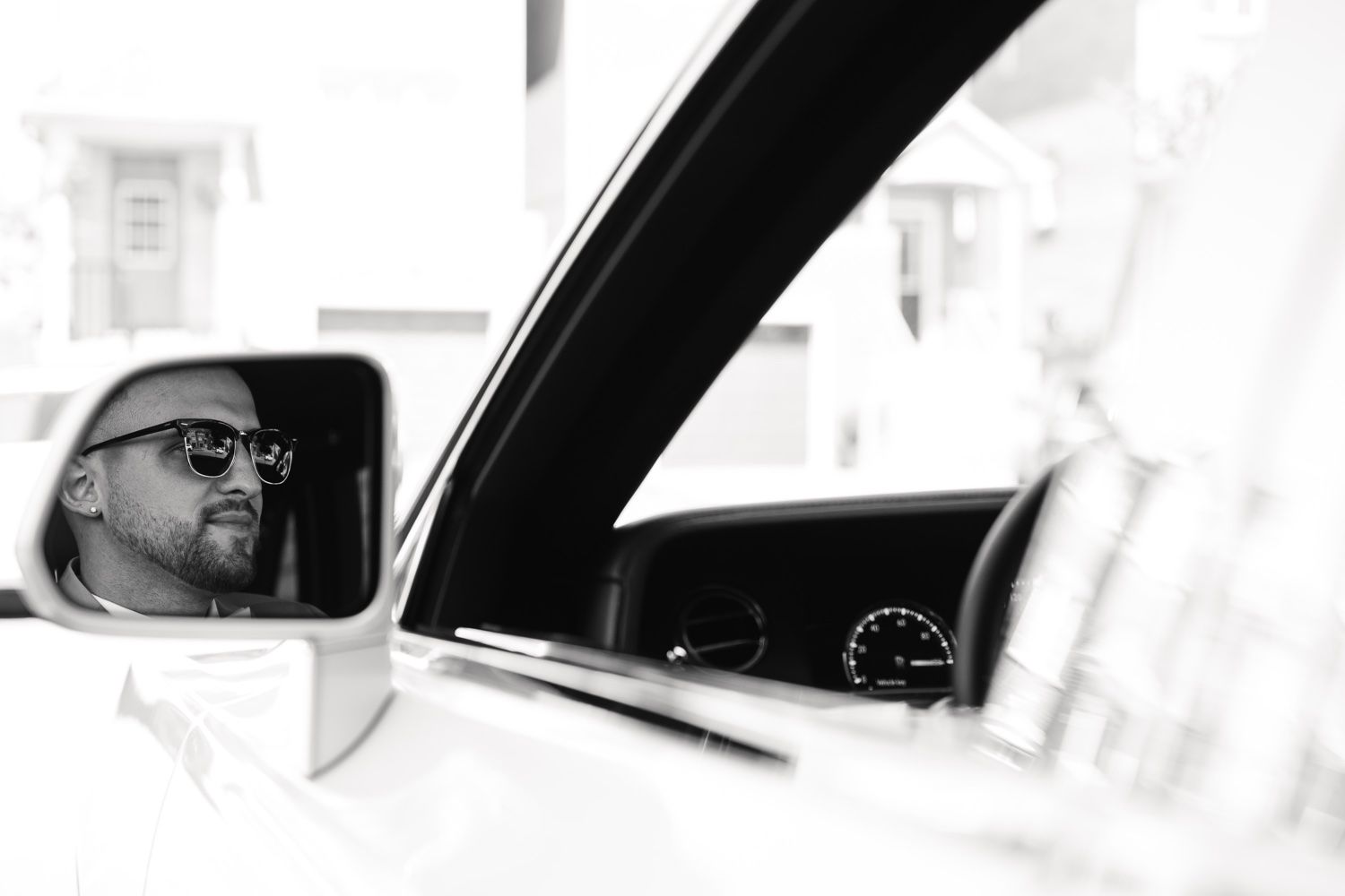 Artistic black and white reflection shot of groom in car mirror wearing sunglasses.