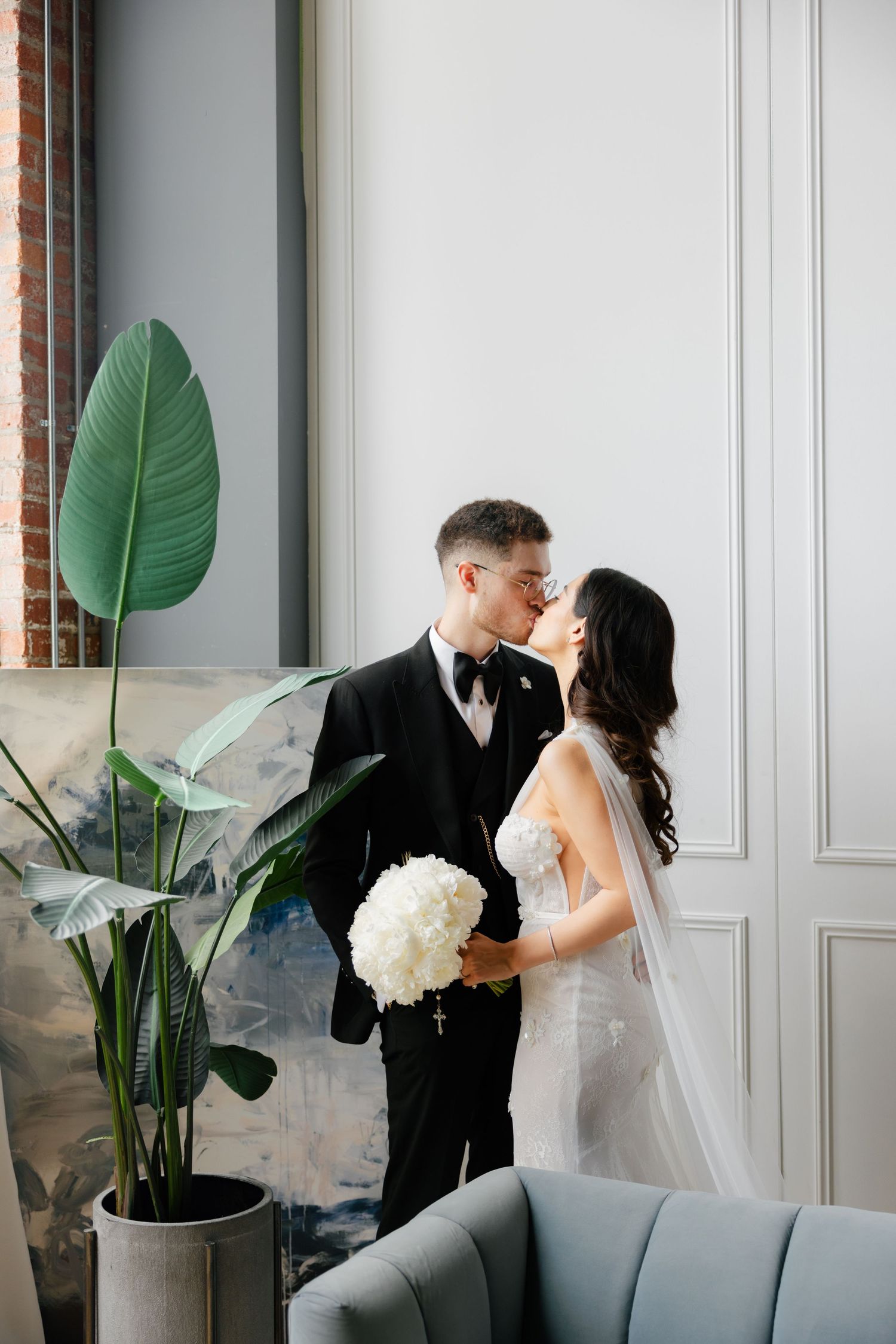 Wedding couple poses beside large potted plant in modern minimalist setting.