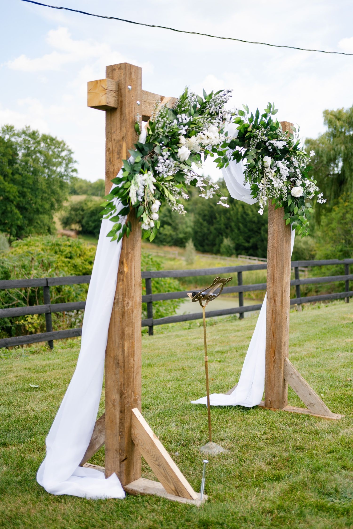 Rustic wooden wedding arch decorated with white flowers, greenery and flowing fabric stands in a garden setting.