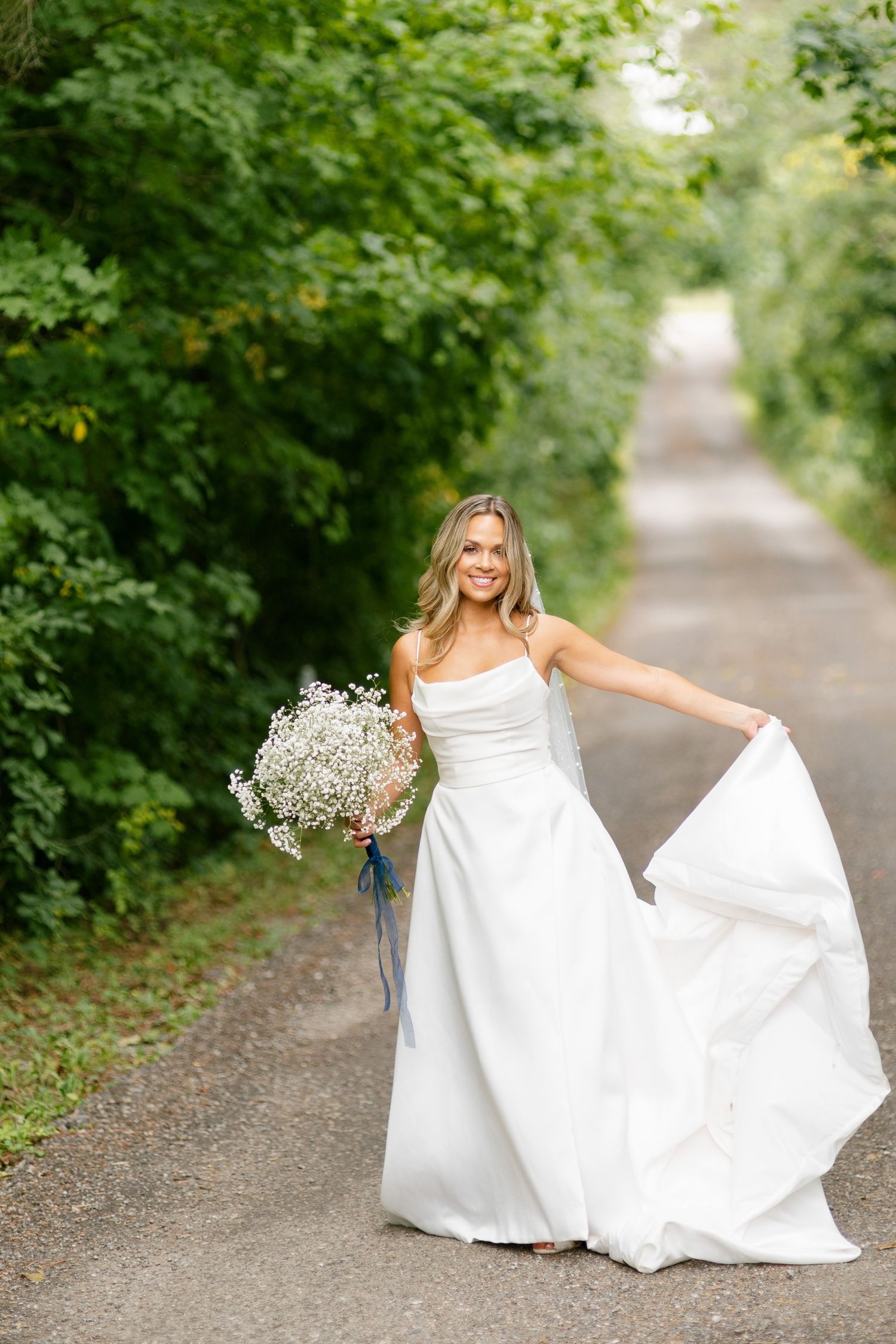 Bride in flowing white gown twirls with baby's breath bouquet on tree-lined path.