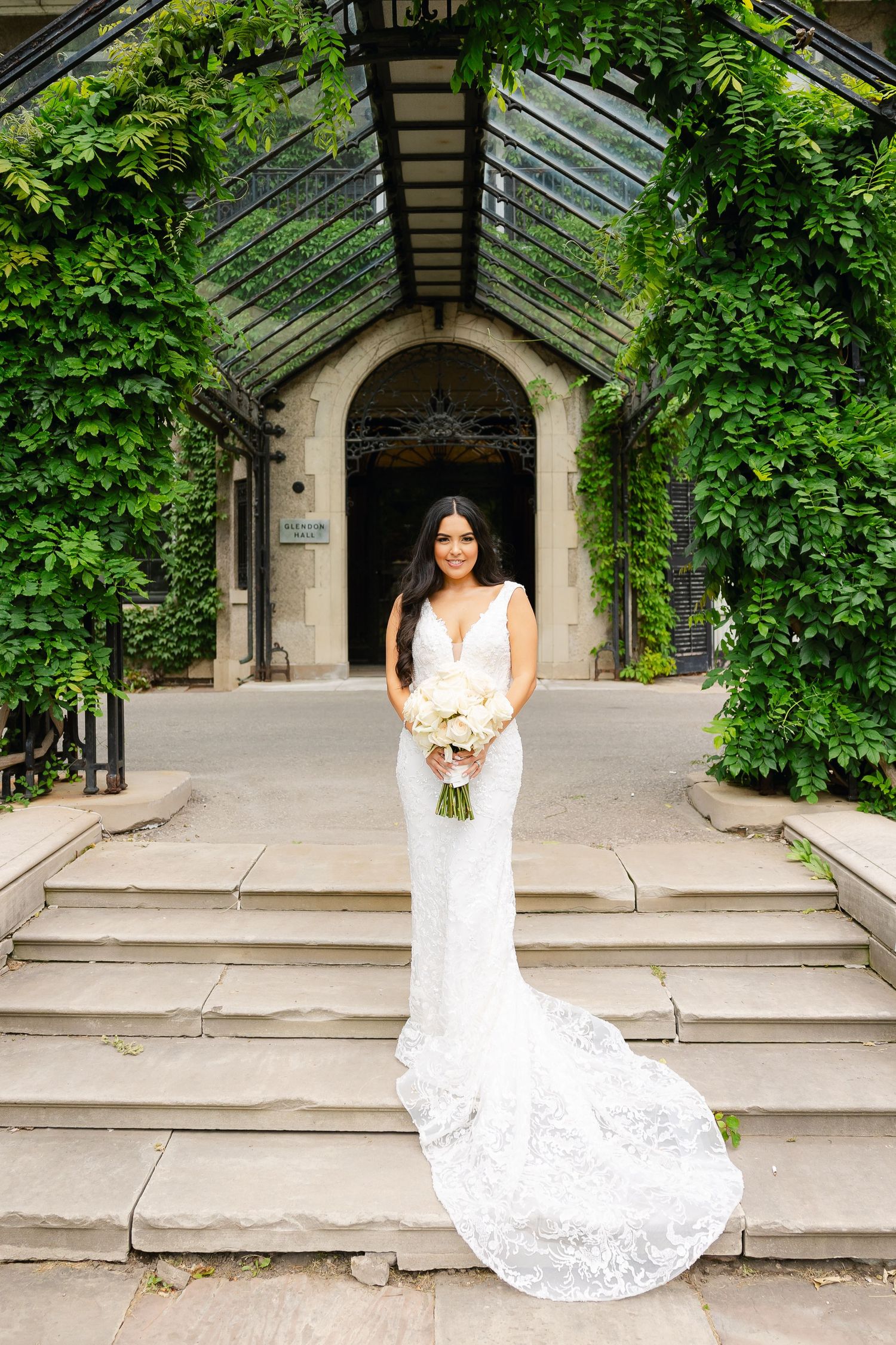 Elegant bride in lace wedding gown with long train poses on stone steps framed by ivy-covered greenhouse entrance.