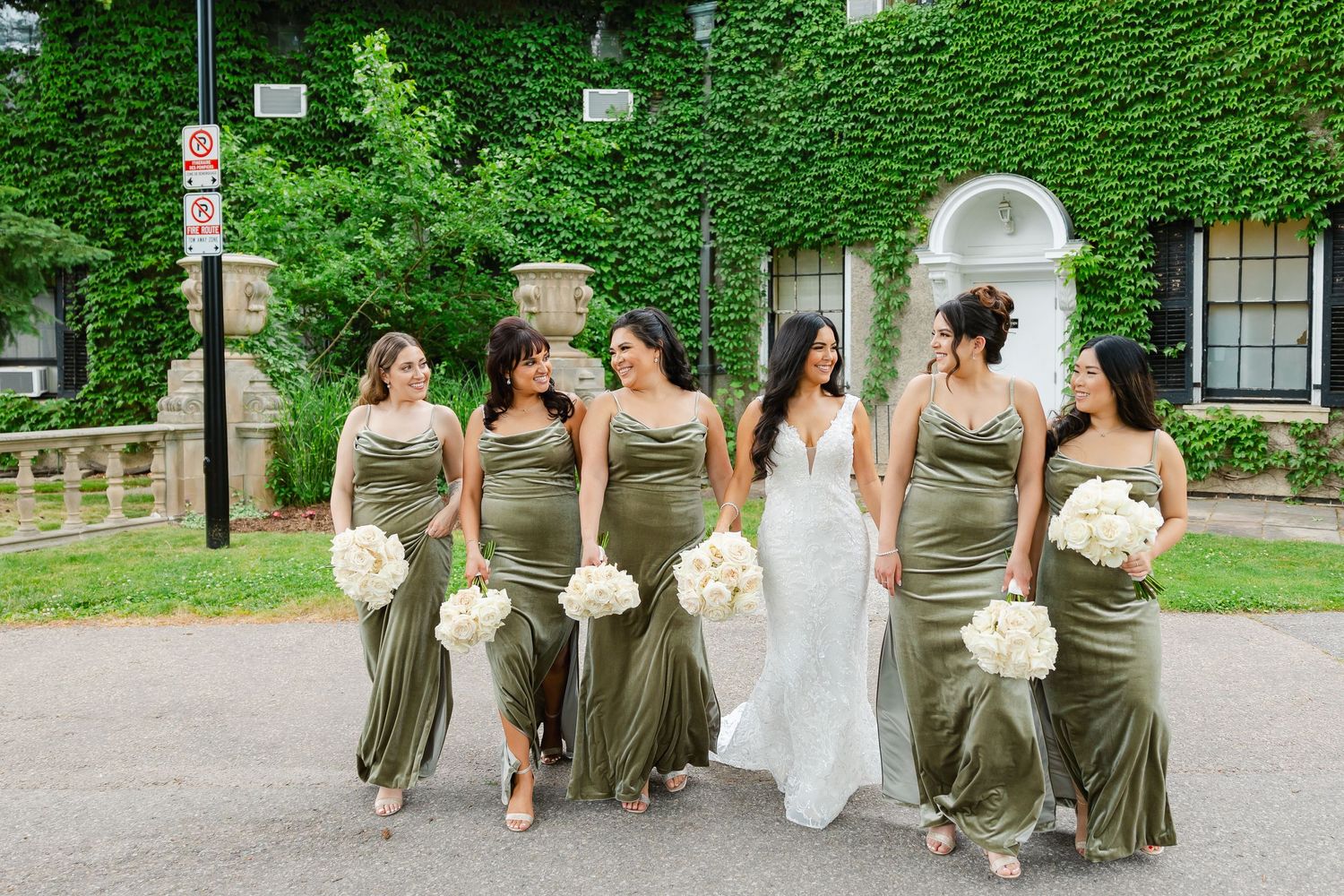 Bridal party in sage dresses walks together outside ivy-covered building.