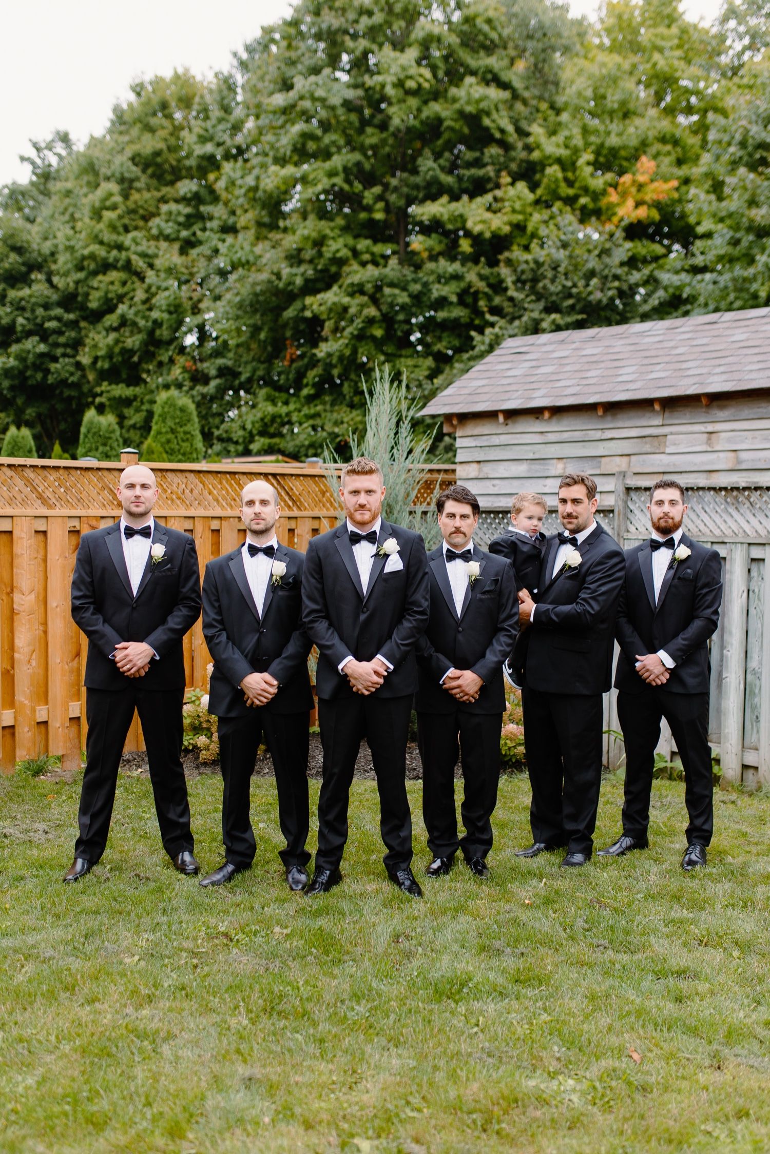 Formal wedding party in black tuxedos pose together in garden setting with wooden fence and shed.