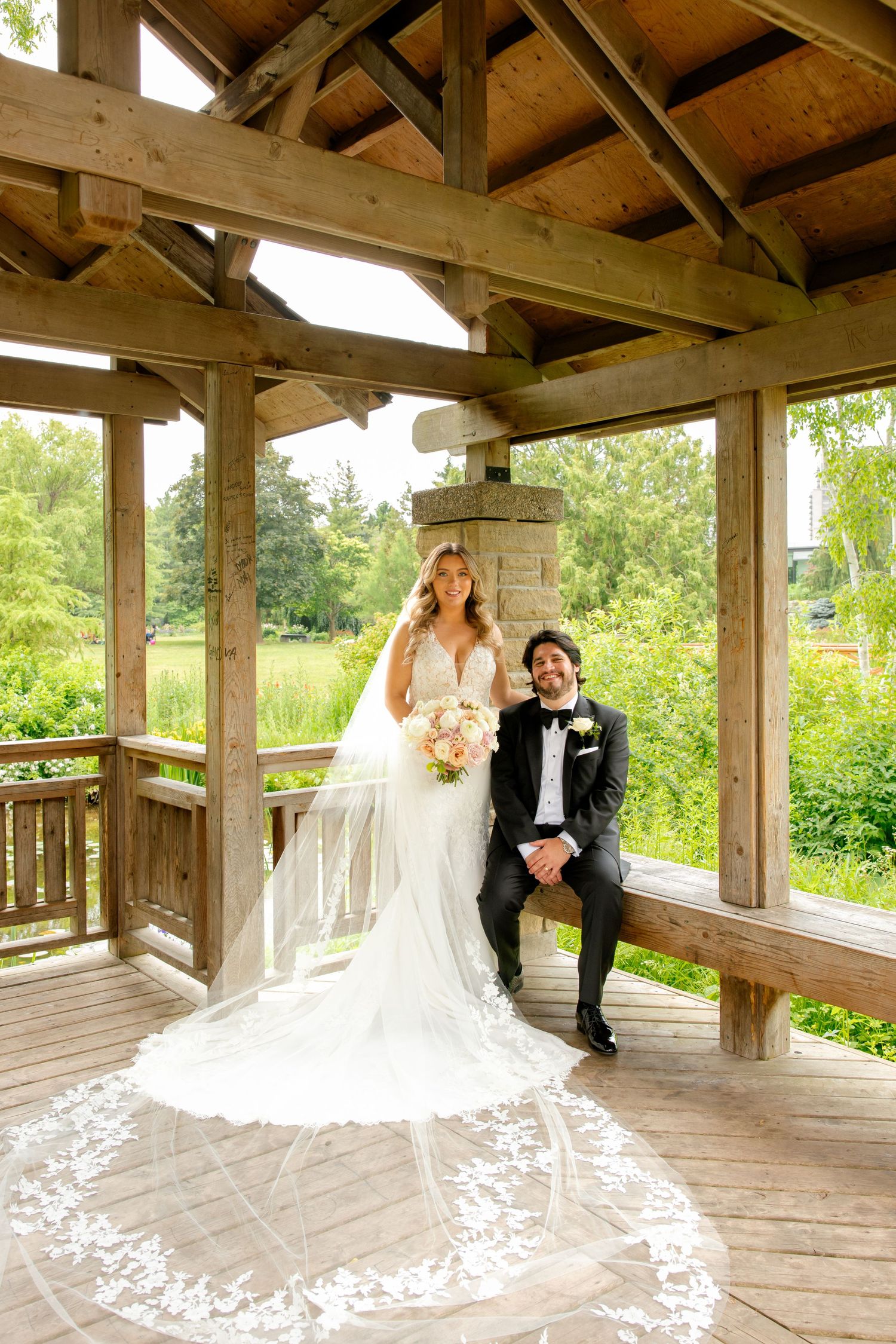 Wedding couple poses in a rustic wooden pavilion with dramatic cathedral veil spread out.
