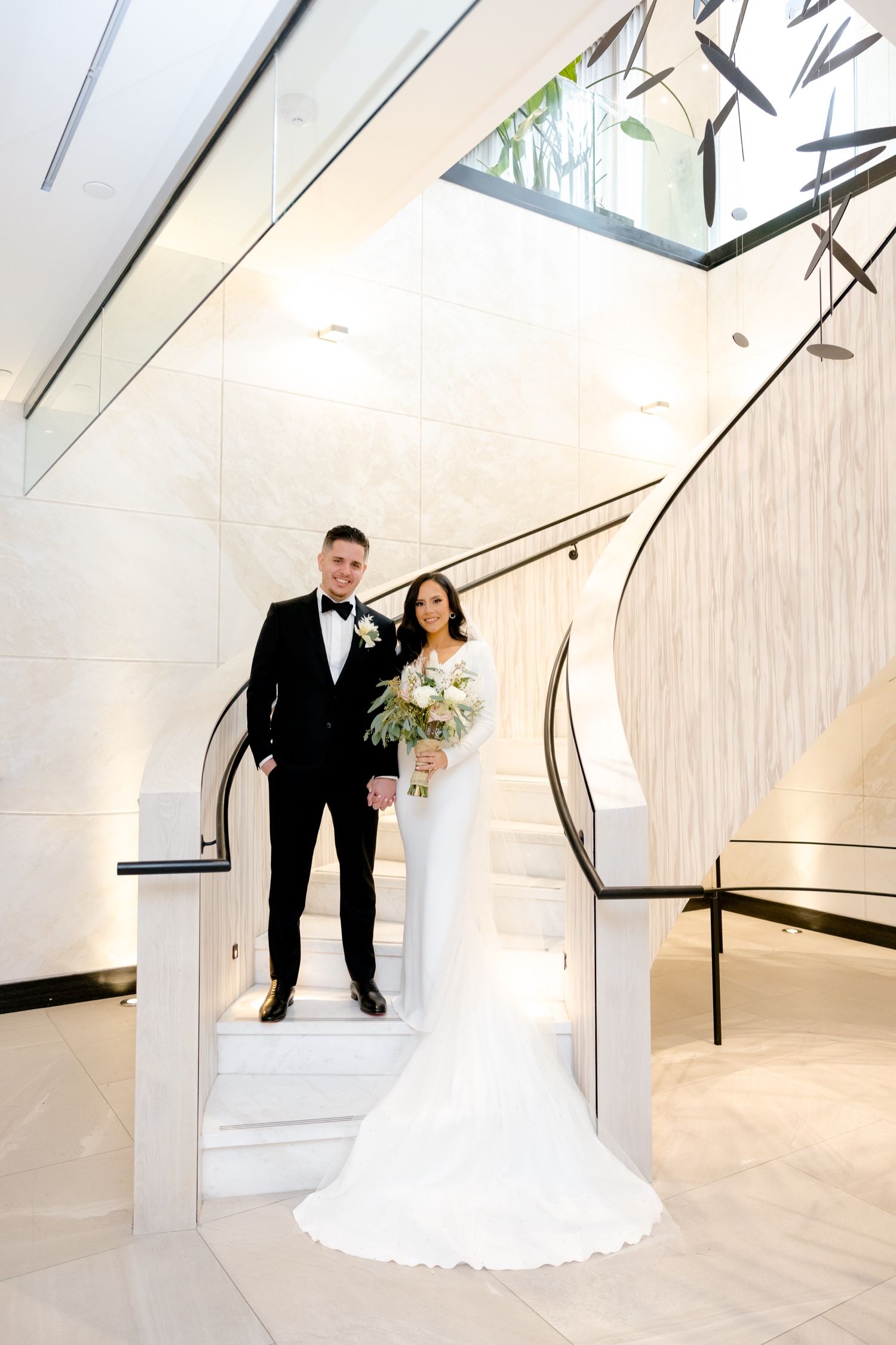 A full-length portrait of newlyweds standing on a modern white curved staircase.
