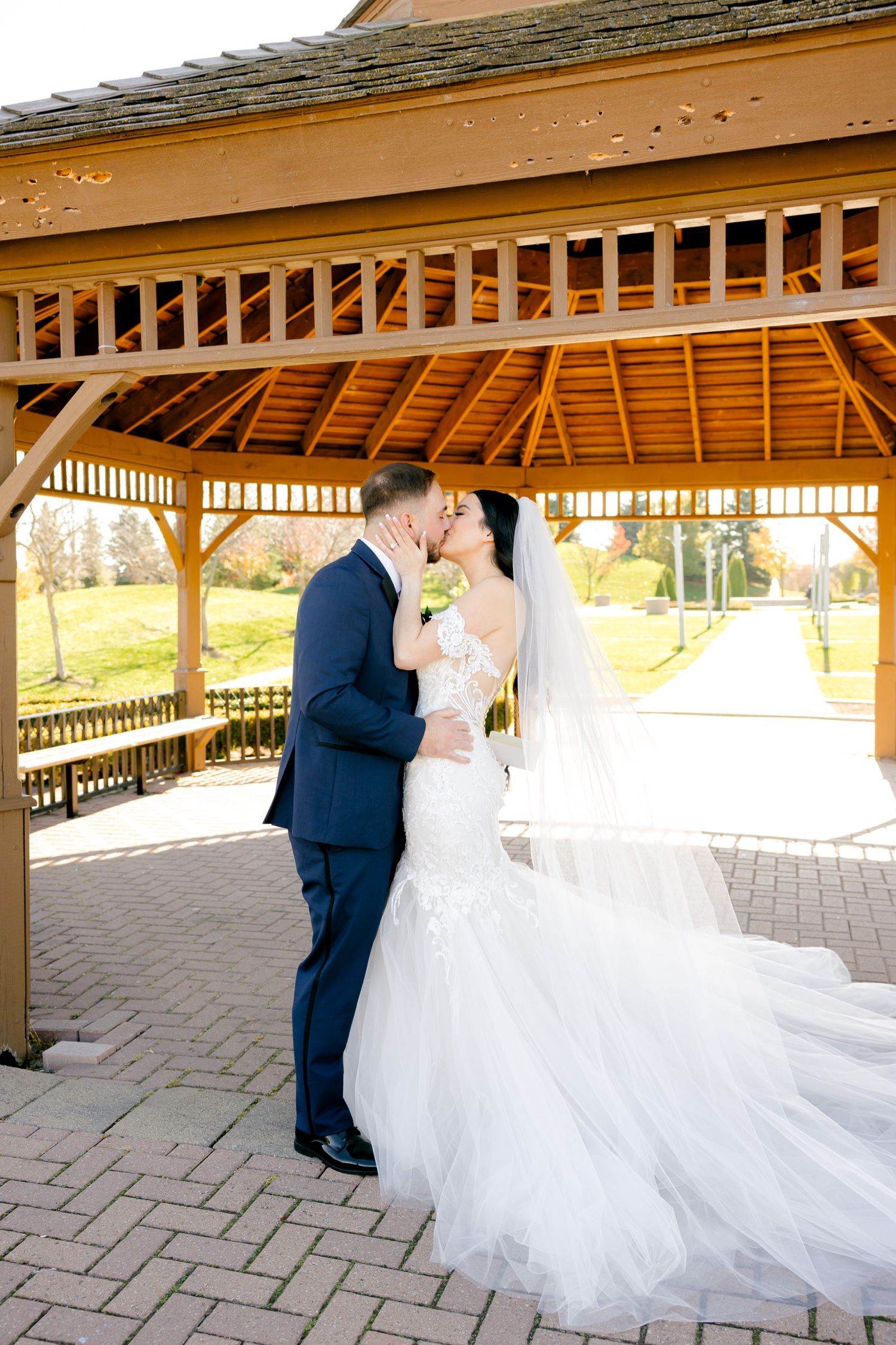 Beautiful wedding couple kisses under wooden gazebo with flowing white dress and veil.