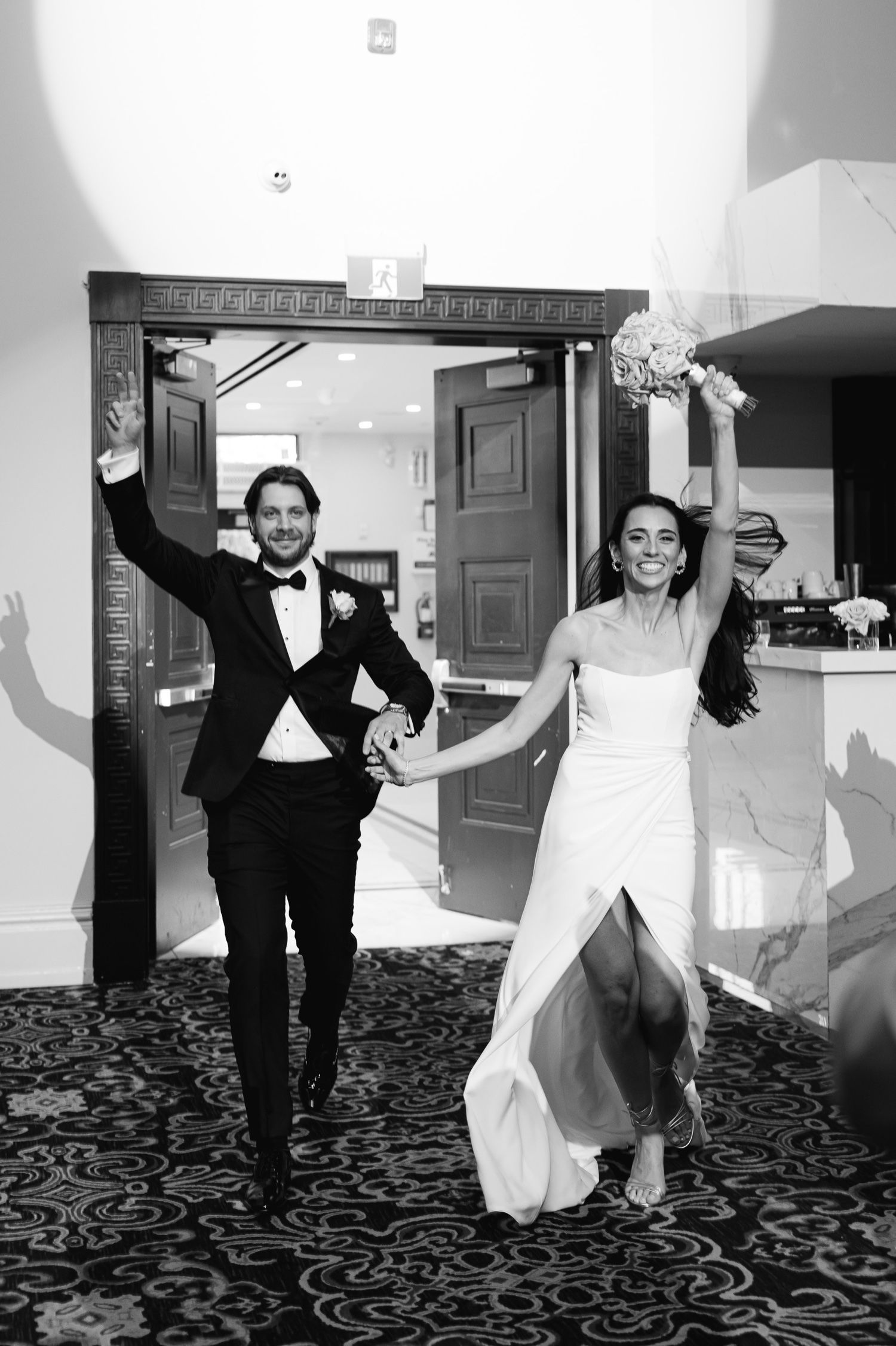 Black and white photo of newlyweds making joyful entrance through ornate doorway.