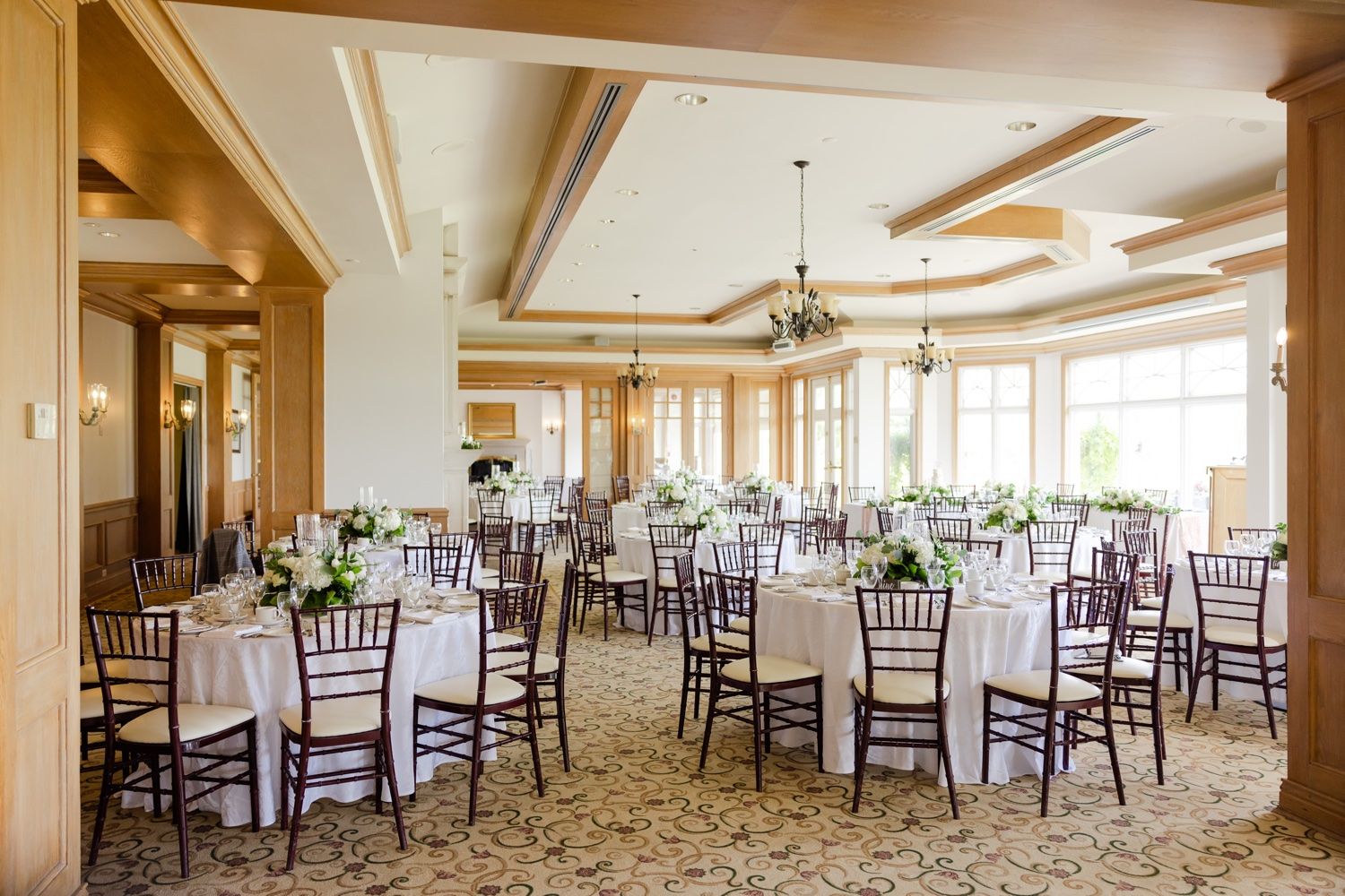 Spacious wedding reception venue with wooden beam ceiling and dark chiavari chairs at round tables.